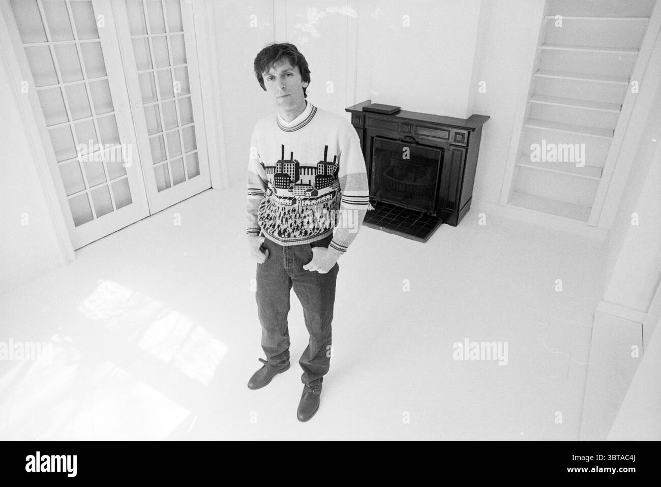 Herr Faasen in Bloemendaal Galerie Bloemendaal, Whizgle News, Dutch Desk, Niederlande, 1950 - 2000 am 24-04-1992. Diese Themen sind in der Abbildung dargestellt. Die Szene zeigt ein Individuum, das selbstbewusst in einem minimalistischen, überwiegend weißen Raum steht. Die Wände und der Boden sind schlank und hell, was ein geräumiges und luftiges Gefühl schafft. Das Sonnenlicht filtert ein, wirft weiche Schatten und verleiht der Kühle der weißen Umgebung eine sanfte Wärme. Die Person trägt einen markanten Pullover, der einen optisch auffälligen Kontrast zur hellen Umgebung bietet. Der Pullover zeigt ein grafisches Design Stockfoto