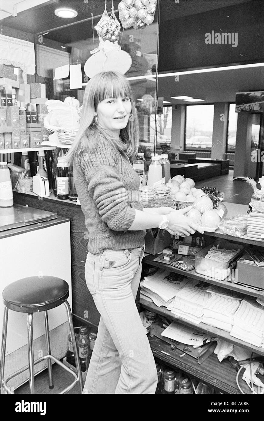 Blumenladen Lebensmittelgeschäft Redaktion Monkfish Friseur (Zeeweg Krankenhaus) Zeeweg Krankenhaus IJmuiden Zeeweg Niederlande, Whizgle News, Dutch Desk, Niederlande, 1950 - 2000 auf 01-04-1977. Diese Themen werden in der Abbildung angezeigt. In einem gemütlichen, geschäftigen Geschäft steht eine junge Frau hinter einer Theke voller verschiedener Gegenstände. Sie hat lange, glatte Haare und ein warmes Lächeln, das die einladende Atmosphäre noch verstärkt. In einem bequemen Strickpullover und einer hoch geschnittenen Jeans spiegelt ihre Kleidung eine lässige, aber stilvolle Ästhetik wider. Die Theke ist mit einer Reihe von Waren versehen, einschließlich einer ordentlichen Stockfoto