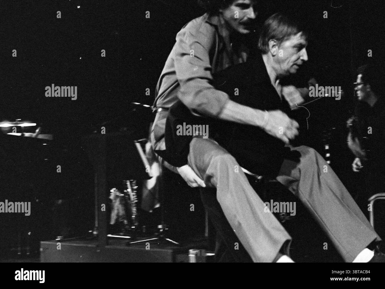 Robert Long und Leen Jongewaard Robert Long, Whizgle News, Dutch Desk, Niederlande, 1950 - 2000 am 01-11-1979. Das Bild enthält diese Themen. In einer lebendigen Bühnensituation stehen zwei Männer im Mittelpunkt der Szene. Der erste Mann, der hinter dem zweiten steht, scheint ihn hochzuheben und eine dynamische Aktion zu kreieren, die Aufmerksamkeit erregt. Der Mann, der angehoben wird, hat einen ernsten Ausdruck, der im Kontrast zur spielerischen Natur des Augenblicks steht. Der Hintergrund ist mit Schatten und sanfter Beleuchtung gefüllt, was auf eine intime Atmosphäre hindeutet, die typisch für eine Vorstellung oder einen Proberaum ist. Es gibt Hinweise auf Musik Stockfoto