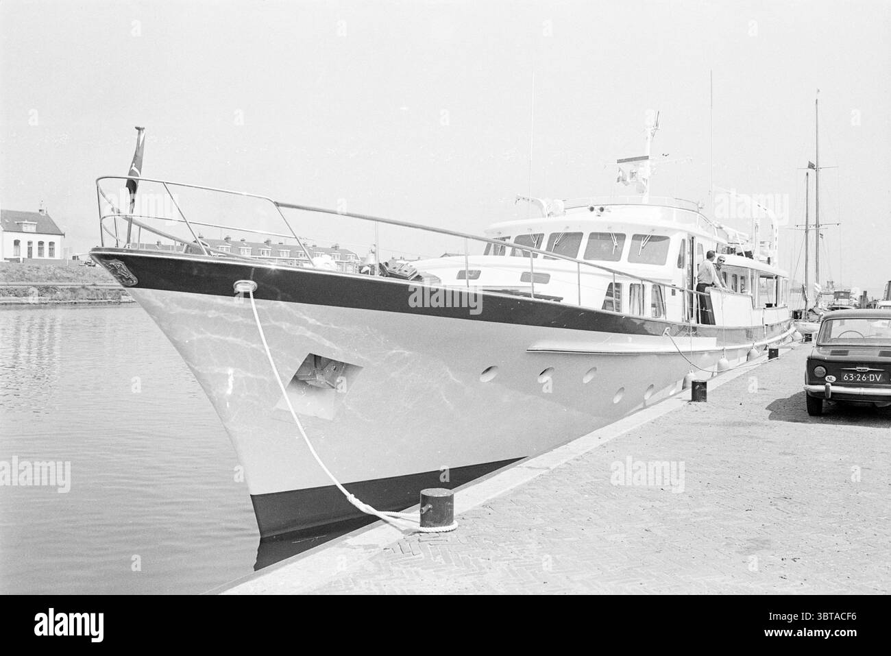 Friedensschiff in IJmuiden Ships, Whizgle News, Dutch Desk, Niederlande, 1950 - 2000 am 13-06-1969. Das Bild enthält diese Themen. Die Szene zeigt ein großes Freizeitboot, das entlang einer ruhigen Wasserstraße vor Anker liegt. Das Schiff hat ein schlankes, stromlinienförmiges Design, das hauptsächlich in einer hellen Farbe mit kontrastfarbenen dunklen Akzenten entlang des Rumpfes gemalt ist. Die Fenster sind groß und gut definiert und spiegeln den geräumigen Innenraum wider. Mehrere Fahnen flattern sanft vom Mast des Bootes und fügen einen Hauch von Farbe gegen die ansonsten gedämpften Töne hinzu. Im Hintergrund umfasst eine malerische Anlage kleine Gebäude entlang der WA Stockfoto