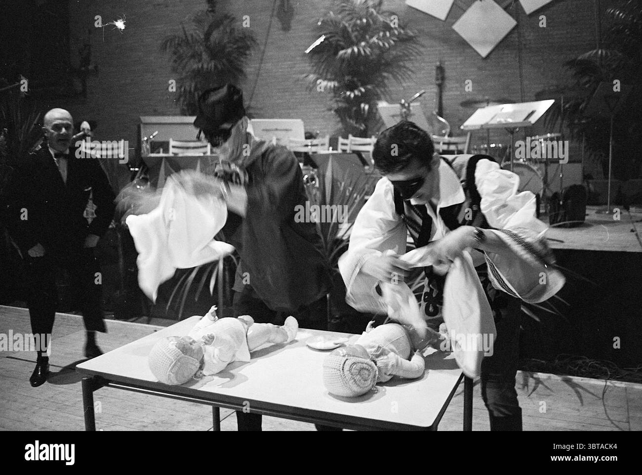 Bunte Abend HST Hochofen Schachturnier, Whizgle News, Dutch Desk, Niederlande, 1950 - 2000 am 25-01-1968. Dies sind die Elemente im Bild. In einem schwach beleuchteten Innenbereich spielen zwei Künstler eine lebhafte Szene an einem Tisch, der mit zwei handgefertigten Puppen geschmückt ist. Der Hintergrund deutet auf eine festliche Atmosphäre mit einer subtilen Bühne und mehreren Instrumenten hin, die darauf warten, gespielt zu werden, was darauf hindeutet, dass eine musikalische Aufführung unmittelbar bevorsteht. Die Darsteller in kontrastierenden Kostümen verleihen der Szene eine verspielte Dynamik. Einer trägt einen klassischen Smoking, komplett mit einer Fliege, und scheint Observi zu sein Stockfoto