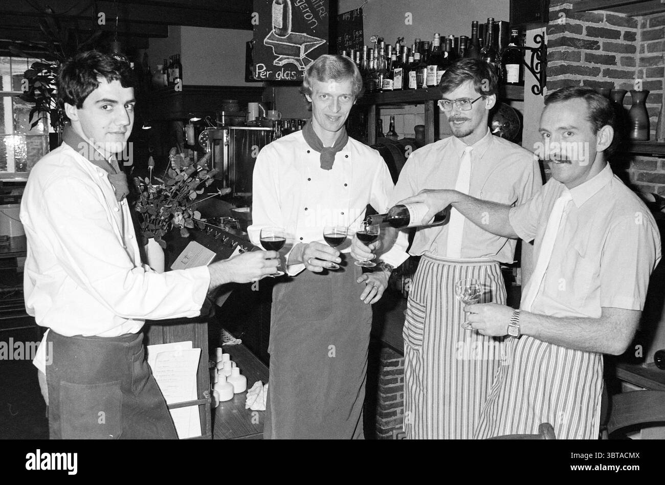 Hans Schouten mit Mitarbeitern des Restaurants Gekroonde Hamer Catering Industry Restaurant, Whizgle News, Dutch Desk, Niederlande, 1950 - 2000 unter 31-10-1983. Die Abbildung zeigt diese Themen. Die Szene fängt einen lebendigen Moment in einem Restaurant mit Wärme und Kameradschaft ein. Vier männliche Server stehen zusammen, die jeweils in knusprigen weißen Hemden und dunklen Schürzen gekleidet sind, was ihre Professionalität und Liebe zum Detail unterstreicht. Der erste Server auf der linken Seite hat kurze, dunkle Haare und zeigt ein höfliches Lächeln, während er ein Glas hält. Neben ihm trägt ein größerer, blonden Kollege ein leichtes Grinsen Stockfoto