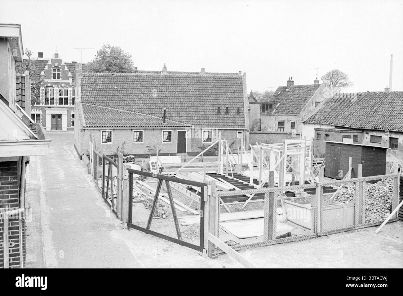 Bauhaus Oud Velsen Construction Construction Pits, Whizgle News, Dutch Desk, Niederlande, 1950 - 2000 am 14-05-1969. Dies sind die Elemente im Bild. Die Szene zeigt ein Baugebiet, eingebettet in eine malerische Wohngegend. Im Vordergrund zeigt ein teilweise erbautes Gebäude Holzrahmen, die auf die Anfänge eines neuen Bauwerks hinweisen. Die Materialien – Hölzer und Versteifungen – sind verstreut, einige sind ordentlich gestapelt, während andere in Verwirrung geraten, was auf laufende Arbeiten hindeutet. Rund um das Gelände befindet sich ein stabiler Holzzaun, der in einem gedämpften Ton gemalt ist und auf eine Grenze hinweist Stockfoto