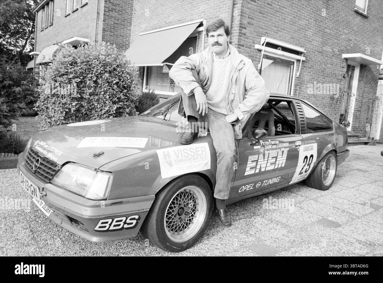 Peter Fontein Rallyefahrer Hillegom, Whizgle News, Dutch Desk, Niederlande, 1950 - 2000 am 22-05-1991. Diese Themen werden in der Abbildung angezeigt. In einer Wohngegend, die von gemauerten Häusern geprägt ist, posiert ein Mann selbstbewusst auf einem Auto, das auf einer kopfsteingepflasterten Einfahrt geparkt ist. Das Fahrzeug ist in leuchtendem Rot lackiert und mit verschiedenen Rennaufklebern versehen, die auf seine modifizierte Natur und seine Leistungsorientierung hinweisen. Der Mann mit einem gut gepflegten Schnurrbart und einer robusten Konstruktion trägt ein lässiges und dennoch stilvolles Ensemble, das einen hellen Pullover und eine dunkle Hose umfasst, die einen Hauch von entspanntem C projiziert Stockfoto
