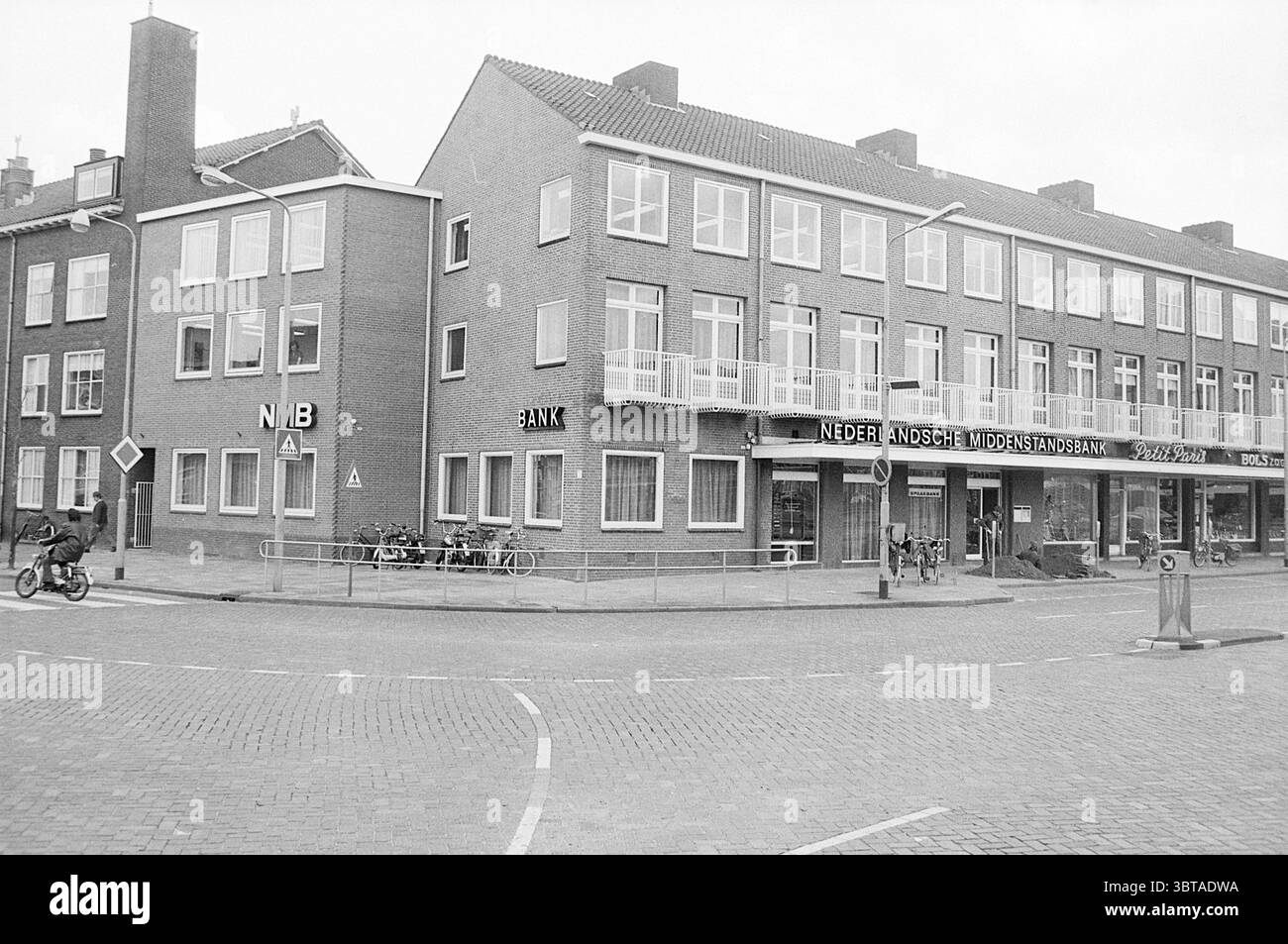 Außenansicht der Netherlandssche Middenstandsbank IJmuiden Plein 1945 The Netherlands, Whizgle News, Dutch Desk, Niederlande, 1950 - 2000 am 19-03-1974. Das Bild enthält diese Themen. Die Szene fängt eine Straßenecke in einer urbanen Umgebung ein, die durch eine Mischung aus architektonischen Stilen gekennzeichnet ist. Auf der einen Seite befindet sich ein dreistöckiges Gebäude mit einem flachen Dach, das mit mehreren Fenstern geschmückt ist, die ein Gefühl der Symmetrie widerspiegeln. Das Gebäude verfügt über eine helle Ziegelfassade, die durch die dunkleren Fensterrahmen leicht abgesetzt wird. Direkt daneben befindet sich ein weiteres Gebäude mit kontrastierendem Design – ein kleineres, zweistöckiges Gebäude – Boa Stockfoto