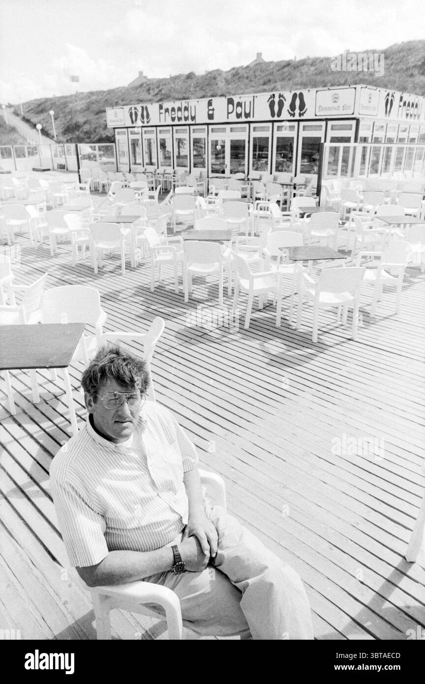 Besitzer Freddy and Paul Meerendonk Cafe Beach and Beaches, Whizgle News, Dutch Desk, Niederlande, 1950 - 2000 auf 31-05-1989. Das Bild enthält diese Themen. Die Szene fängt eine ruhige Umgebung im Freien mit einer weitläufigen Holzterrasse ein, die sich zu einer Reihe weißer Stühle und Tische erstreckt, die ordentlich angeordnet sind. Die Atmosphäre lässt einen Küstenort nahe einem Strand vermuten, was durch die zwanglose, entspannte Atmosphäre verdeutlicht wird. Im Vordergrund sitzt ein Mann mittleren Alters bequem auf einem weißen Plastikstuhl und trägt ein helles kurzärmeliges Hemd. Sein Ausdruck ist kontemplativ, vielleicht reflektierend, Stockfoto