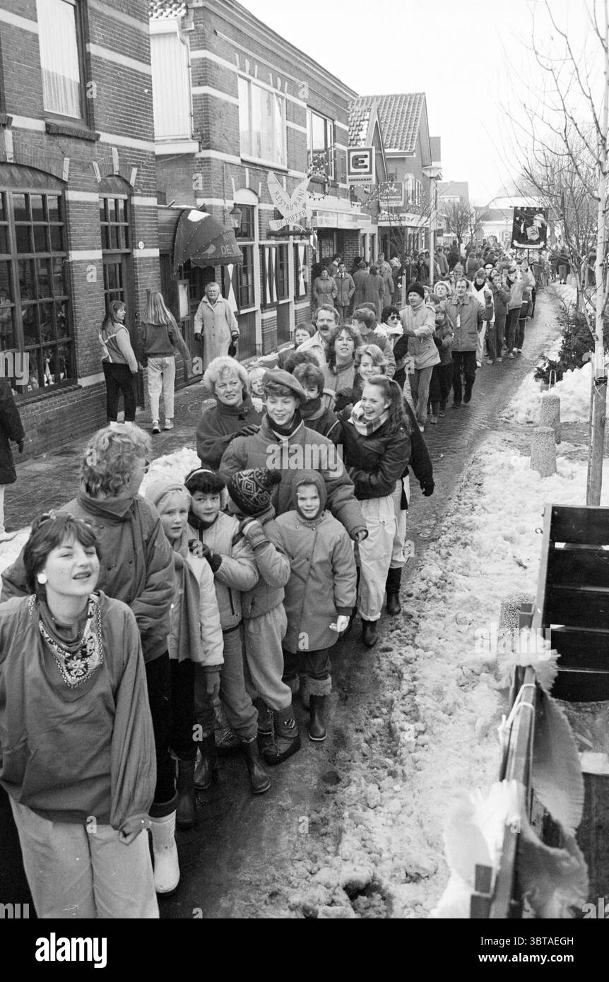 Lange Polonaise durch Noordwijkerhout Noordwijkerhout Rekordversuche und Verbesserungen Noordwijkerhout, Whizgle News, Dutch Desk, Niederlande, 1950 - 2000 am 12-01-1985. Das Bild enthält diese Themen. Die Szene zeigt eine lebhafte Winterfeier in einer urbanen Umgebung, bei der eine Vielzahl von Individuen eine einzelne Aktenlinie bilden, während sie auf einer schneebedeckten Straße spazieren. Die Architektur der Gebäude, die die Straße flankieren, ist typisch für malerische Städte, mit roten Ziegelfassaden und dekorativen Elementen, die eine festliche Atmosphäre vermitteln. Die Menschen in der Prozession zeigen eine Reihe von Emotionen Stockfoto