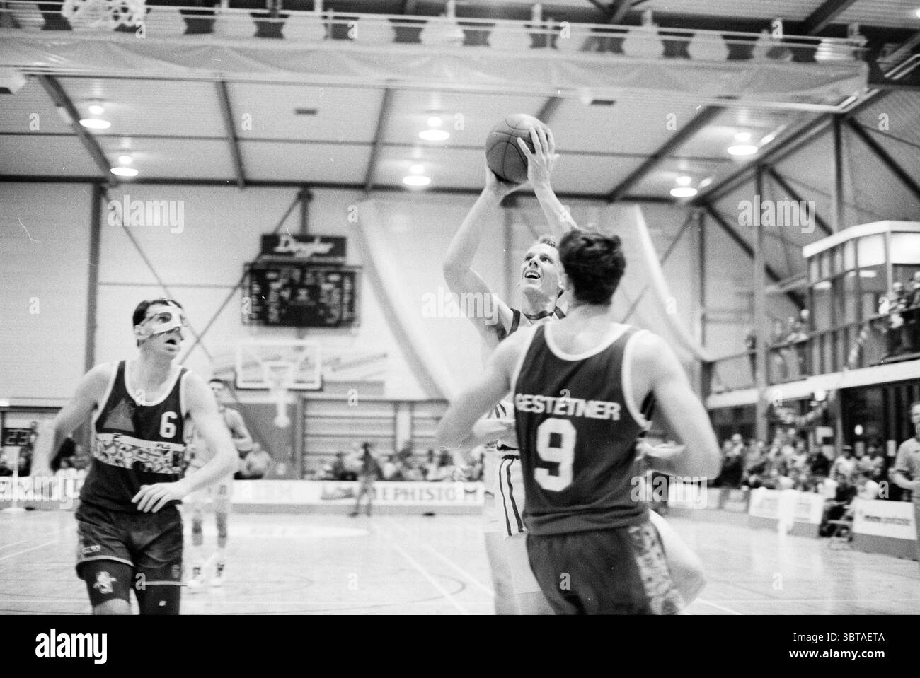 Bestmate - Nashua, Whizgle News, Dutch Desk, Niederlande, 1950 - 2000 am 22-02-1991. Diese Themen werden in der Abbildung angezeigt. Die Szene fängt einen intensiven Moment während eines Basketballspiels in einer geräumigen Sporthalle ein. Der polierte Holzboden des Hofes bildet einen warmen Kontrast zu den kühlen Grautönen der umgebenden Struktur und ihrer metallischen Komponenten. Im Vordergrund befindet sich ein Spieler mitten im Sprung und greift mit Entschlossenheit nach dem Basketball. Sein sportlicher Aufbau und sein konzentrierter Ausdruck betonen die Intensität des Augenblicks. Er trägt ein helles Trikot mit vertikalem Streifen Stockfoto