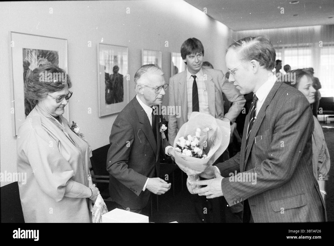 Empfang Herr Lettinga Reception, Whizgle News, Dutch Desk, Niederlande, 1950 - 2000 am 02-05-1986. Dies sind die Elemente im Bild. In einem hellen, modernen Interieur versammelt sich eine kleine Gruppe von Menschen um eine zentrale Figur, die einen Blumenstrauß hält. Die Szene strahlt ein Gefühl von Feiern und Wärme aus. Die Einzelpersonen sind in formeller Kleidung gekleidet, wobei der Mann die Blumen in einem dunklen Anzug und einer Brille präsentiert, die ein professionelles Aussehen zeigt, während eine ältere Frau in einer hellen Bluse in der Nähe steht, klar zufrieden. Das Zimmer verfügt über ein minimalistisches Design, das durch hervorgehoben wird Stockfoto