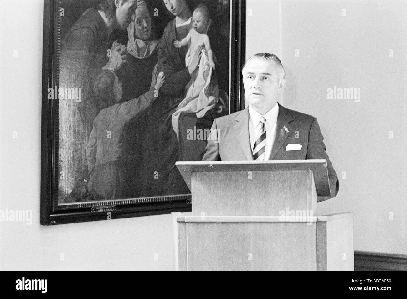 Eröffnung Boerenleenbank Eröffnungen Put in Use, Whizgle News, Dutch Desk, Niederlande, 1950 - 2000 am 24-08-1970. Diese Themen sind in der Abbildung dargestellt. Die Szene verfügt über eine formelle Umgebung, die sich in einem Saal oder einem Raum befindet, der für Tagungen oder Präsentationen konzipiert ist. Ein Mann steht auf einem hölzernen Podium, gekleidet in einem Anzug mit einer Krawatte mit markanten Streifen, die Professionalität zum Ausdruck bringen. Sein Ausdruck ist ernst und konzentriert, was darauf hindeutet, dass er ein Publikum anspricht oder eine Rede hält. Hinter ihm hängt ein großes, gerahmtes Gemälde, das eine Gruppe von Figuren zeigt, die sich in einem intimen, emotionalen ich engagieren Stockfoto