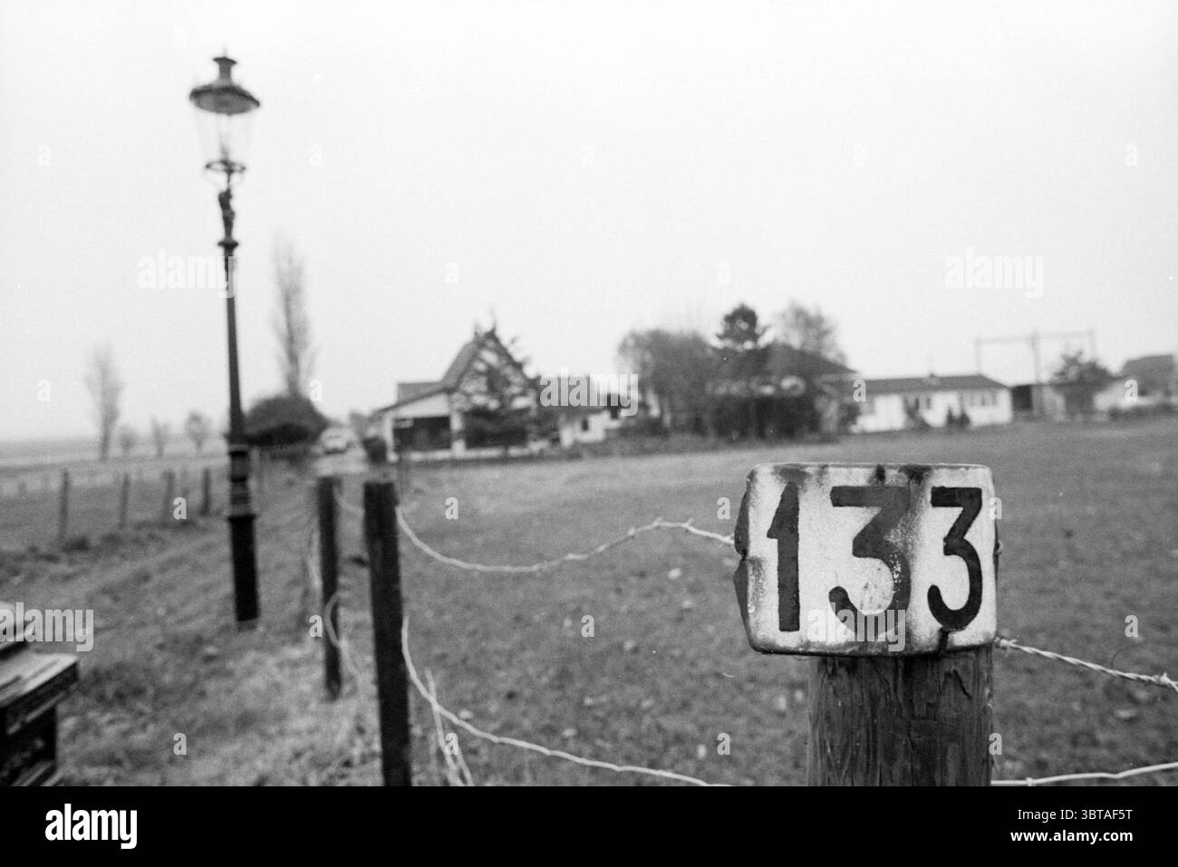 Hausnummern Haarlemmermeer, Whizgle News, Dutch Desk, Niederlande, 1950 - 2000 am 01-11-1993. Dies sind die Themen im Bild. Die Szene präsentiert eine ländliche Landschaft, umgeben von einer gedämpften, monochromatischen Palette, die ein Gefühl von Ruhe und Nostalgie betont. Im Vordergrund verläuft parallel zum Betrachter ein verwitterter Holzzaun, dessen rustikale Pfosten sich mit dem glatten, bewölkten Himmel darüber abheben. Einer der Zaunpfähle trägt die Nummer „133“ in einem auffälligen, aber verblassten Stil, der die Aufmerksamkeit auf die ansonsten dezenten Details der Szene lenkt. Links steht eine altmodische Straßenlaterne Stockfoto