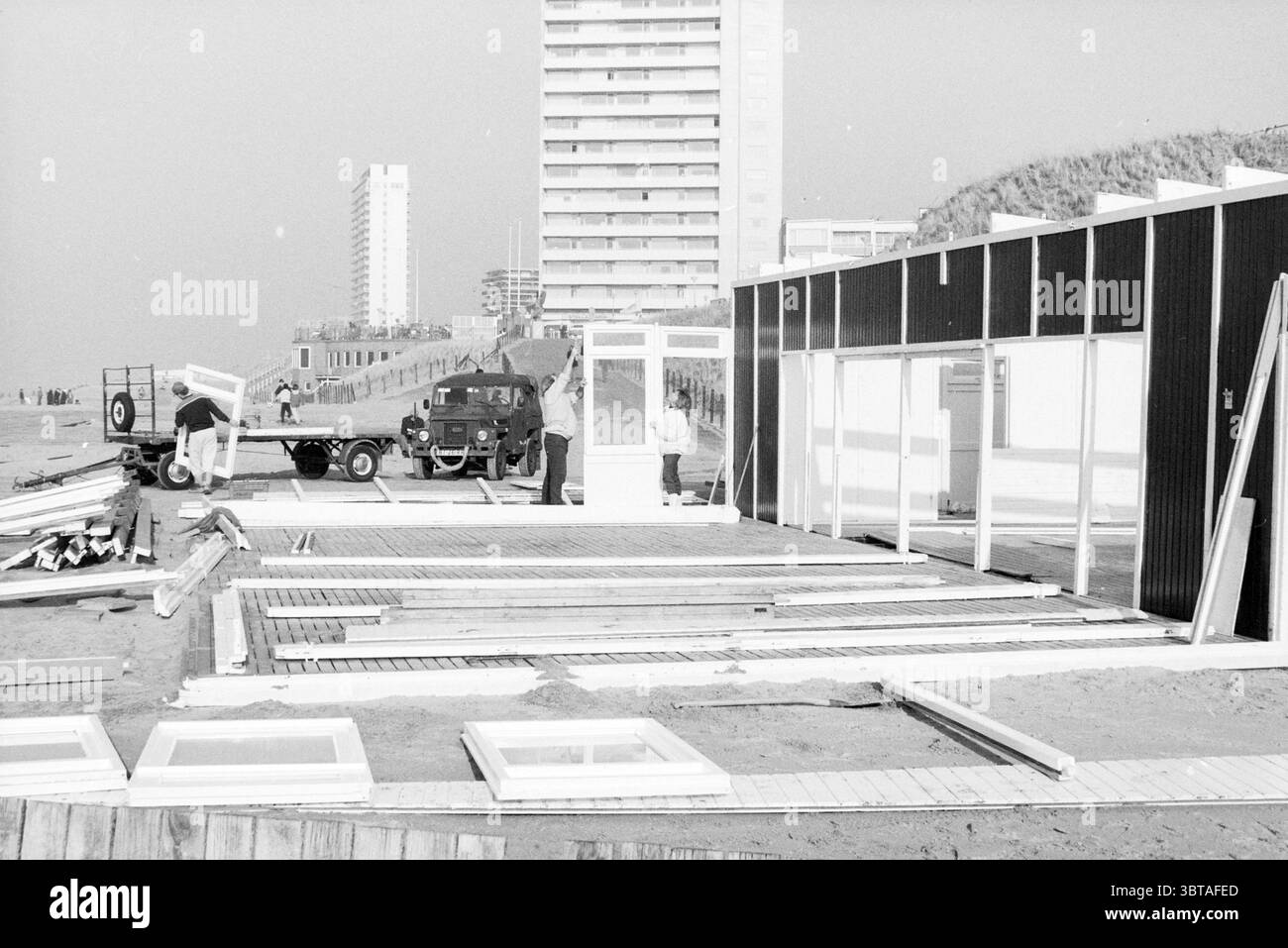 Bau Strandzelte Zandvoort Zandvoort, Whizgle News, Dutch Desk, Niederlande, 1950 - 2000 am 08-02-1989. Das Bild enthält diese Themen. Die Szene zeigt eine Küstenbaustelle, wahrscheinlich in der Nähe eines Strandes. Im Vordergrund befindet sich eine teilweise zusammengesetzte Struktur mit Holzdielen und -Platten, was auf ein laufendes Projekt hindeutet – möglicherweise eine Strandanlage oder einen Kiosk. Die Bretter sind willkürlich angeordnet, einige liegen flach auf dem Boden, während andere auf ihren Kanten gestützt sind, was ein Gefühl von organisiertem Chaos schafft. Links steht ein klassisches Fahrzeug, wahrscheinlich ein Lkw, auf dem geparkt wird Stockfoto
