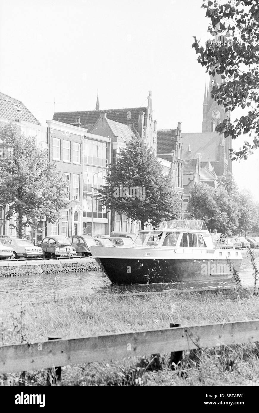 Sun Report in Haarlem Report Summer Pictures Haarlem the Netherlands, Whizgle News, Dutch Desk, Niederlande, 1950 - 2000 auf 30-08-1979. Das Bild enthält diese Themen. Die Szene zeigt einen ruhigen Wasserweg, der von charmanten historischen Gebäuden umgeben ist. Ein bescheidenes Motorboot driftet sanft auf dem Kanal, dessen weißer Rumpf sich vom dunkleren Wasser abhebt, während sanft reflektierte Wellen einen impressionistischen Effekt erzeugen. Um den Wasserweg herum gibt es malerische Bauten, die eine Mischung aus architektonischen Stilen mit Giebeldächern und großen Fenstern zeigen. Sie sind überwiegend in dezenten, gedämpften Tönen, die andeuten Stockfoto