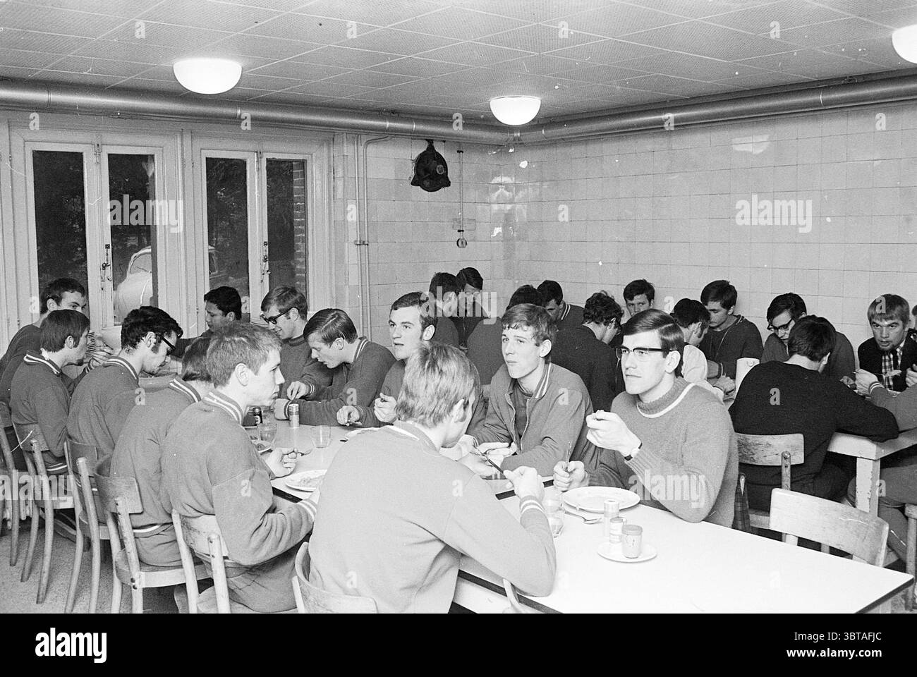 Bericht CIOS C.I.O.S., Whizgle News, Dutch Desk, Niederlande, 1950 - 2000 am 06-12-1968. Das Bild enthält diese Themen. Die Szene zeigt einen Gemeinschaftsbereich mit jungen Männern, die an langen rechteckigen Tischen sitzen. Das Ambiente ist insgesamt etwas gedämpft, das durch die gedämpften Schwarz-weiß-Töne vermittelt wird. Die Gäste sind lässig in Pullover gekleidet, was ein Gefühl der Gleichmäßigkeit schafft. Ihre Ausdrucksformen reichen von Konzentration bis hin zu zwanglosen Gesprächen, was eine Mischung aus Interaktion mit ihren Mahlzeiten und miteinander suggeriert. Teller und Schüsseln sind über die Tische verteilt, was auf einen Anteil hinweist Stockfoto