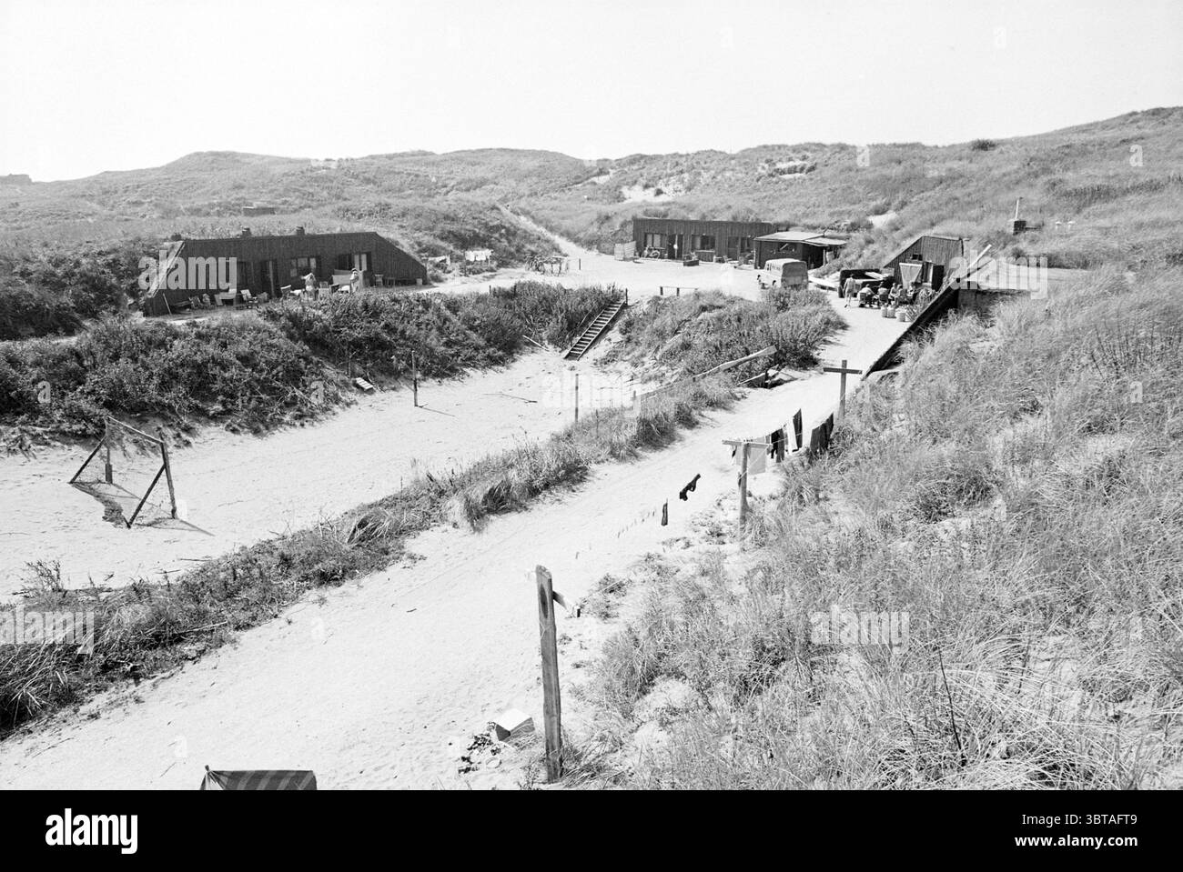 t Schuitegat (Bunker in den Dünen) Zandvoort Bunkers Dunes Zandvoort Zandvoort, Whizgle News, Dutch Desk, Niederlande, 1950 - 2000 am 29-08-1979. Die Abbildung zeigt diese Themen. Die Szene zeigt einen Sandbereich, der von sanften Dünen umgeben ist, die mit hohen Gräsern bedeckt sind. Zwei bescheidene, rustikale Bauten mit Holzwänden und schrägen Dächern stehen in der Mitte, was auf einen Gebrauchszweck hindeutet. Sie wirken verwittert und deuten auf eine lange Exposition gegenüber den Elementen hin. Ein Pfad schlängelt sich durch das sandige Gelände, der vom Vordergrund zu den Bauwerken führt und zu Erkundungen einlädt. Auf beiden Seiten des Weges, Büschel von Stockfoto