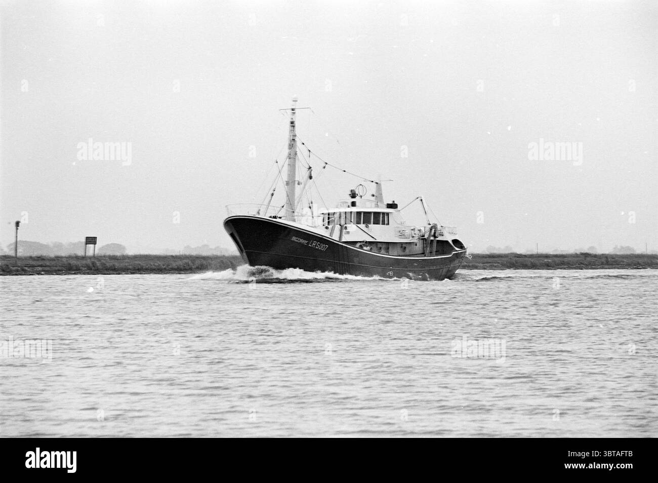 Bigorre in Noordzeekanaal Haarlemse Scheepsbouw Mij, Whizgle News, Dutch Desk, Niederlande, 1950 - 2000 am 20.06.1963. Das Bild enthält diese Themen. Die Szene fängt einen ruhigen grauen Tag auf dem Wasser ein, an dem ein robustes Fischereischiff durch ruhiges Wasser gleitet. Das Boot ist in tiefen Schwarz-weiß-Tönen lackiert und verfügt über eine markante Kabine mit verschiedenen funktionalen Strukturen und Ausrüstungen, die typischerweise für den Fischfang bestimmt sind. Der Rumpf bildet einen subtilen Kontrast zum gedämpften Hintergrund. Um das Schiff herum kräuselt sich das Wasser sanft und erzeugt sanfte Wellen, die die Motive des Bootes widerspiegeln Stockfoto