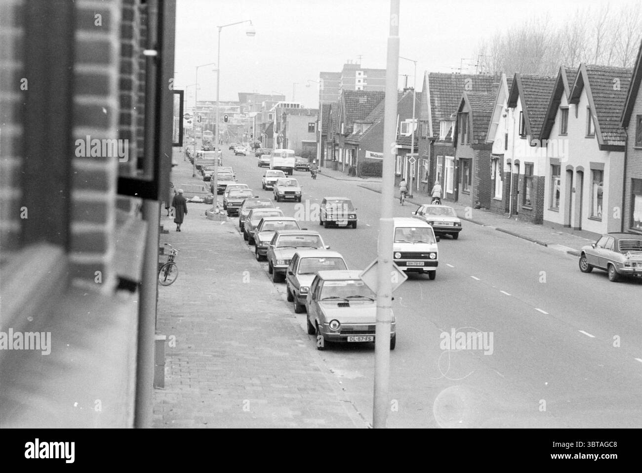 Übersicht Alkmaarseweg Beverwijk Übersicht Straßen Straßenbau Straßenbau Straßenbepflanzung Beverwijk Alkmaarseweg Niederlande, Whizgle News, Dutch Desk, Niederlande, 1950 - 2000 am 08-11-1983. Dies sind die Themen im Bild. Die Szene fängt eine beschauliche urbane Straße ein, gesäumt von Fahrzeugen aus vergangener Zeit, die sich durch ihr kompaktes und klassisches Design auszeichnen. Eine Reihe von Autos, hauptsächlich in gedämpften Grau-, Blau- und Weißtönen, parken am Straßenrand, was auf eine ruhigere Zeit mit weniger Staus hindeutet. Auf der linken Seite befindet sich ein Backsteinbau, dessen Fensterrahmen einen Einblick in die Resi geben Stockfoto