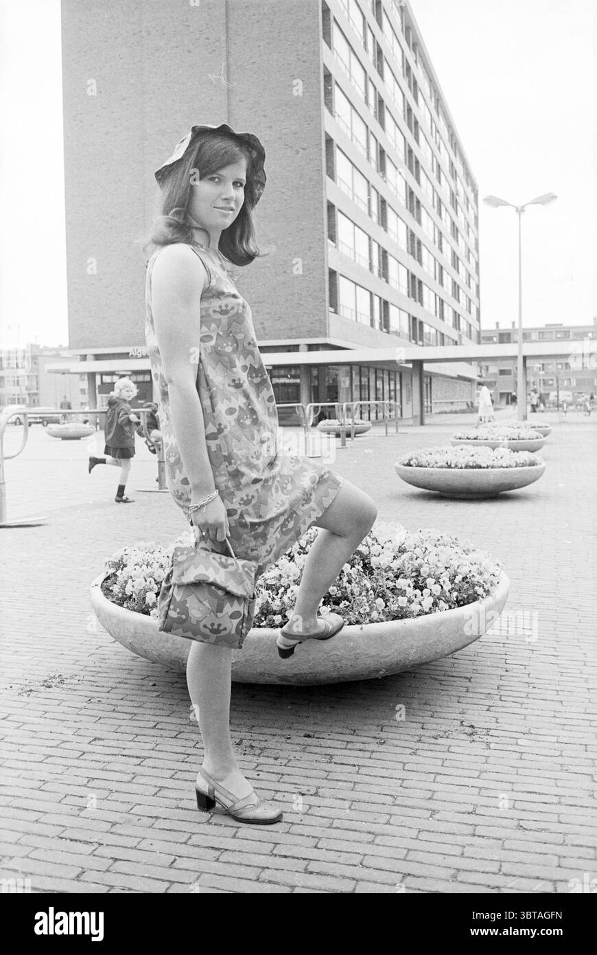Junge Frau posiert im modernen Einkaufszentrum Whizgle News, Dutch Desk, Niederlande, 1950 - 2000 am 05-1968. Dies sind die Elemente im Bild. Die Szene spielt in einer urbanen Umgebung, die durch ein modernes Gebäude im Hintergrund mit einer Reihe von großen Fenstern gekennzeichnet ist. Die Architektur ist unkompliziert, mit einer markanten Fassade, die ein funktionales Design andeutet, das typisch für Bauten aus der Mitte des 20. Jahrhunderts ist. Im Vordergrund steht eine junge Frau selbstbewusst und zeigt ein stilvolles Outfit. Sie trägt ein Kleid mit Blumenmuster und leuchtenden Farben, das eine lebhafte und Jugendliche Atmosphäre vermittelt. Ihr h Stockfoto