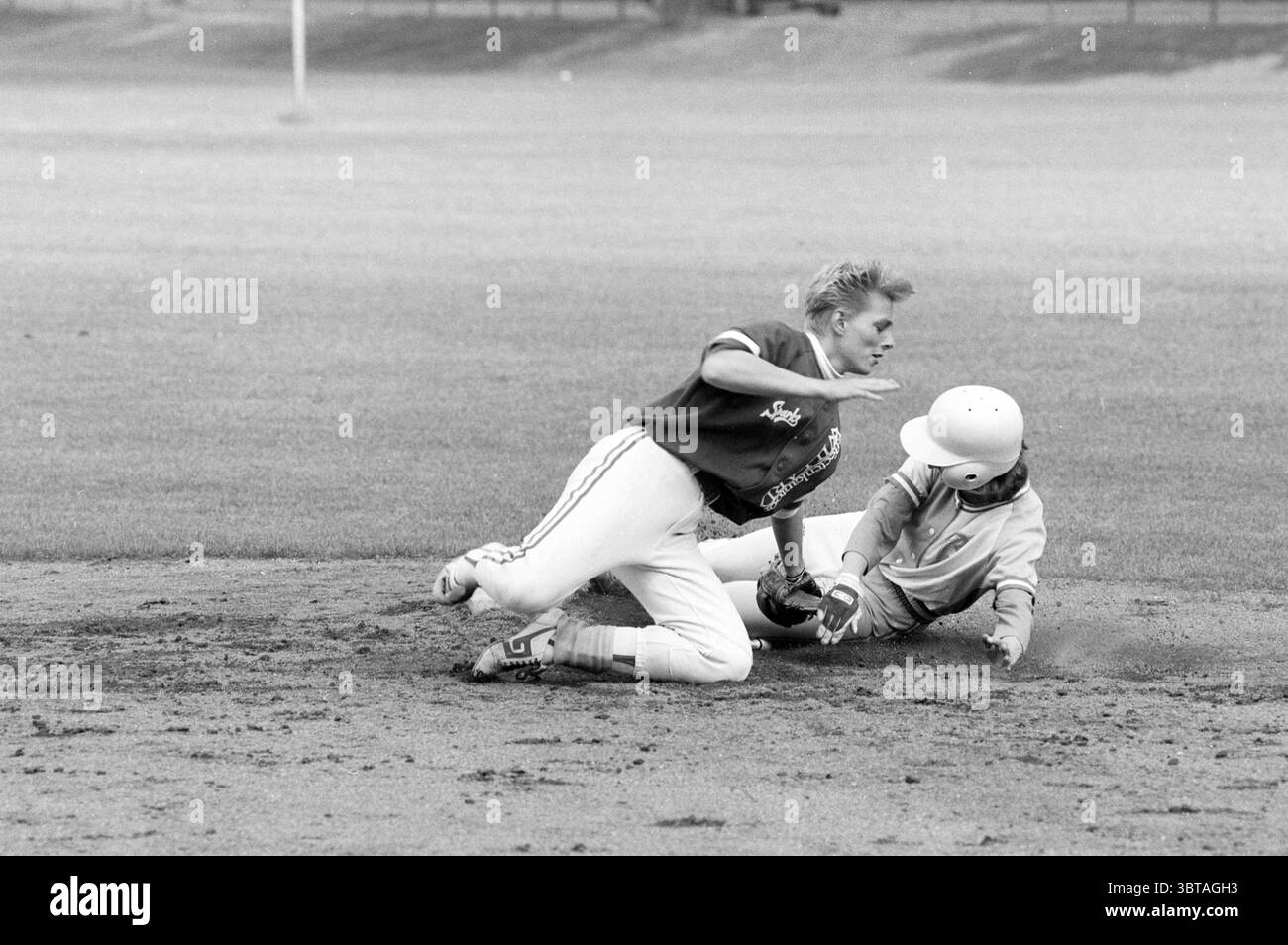 Softball Sparks - Bloemendaal, Whizgle News, Dutch Desk, Niederlande, 1950 - 2000 am 15-06-1991. Das Bild enthält diese Themen. In einer dynamischen Sportszene spielen zwei junge Athleten einen entscheidenden Moment auf einem Baseballdiamanten. Der erste Athlet, der an der Spitze steht, befindet sich in der Mitte der Action und rutscht mit einem entschlossenen Ausdruck in die Basis. In einer kontrastierenden Uniform – einem leichteren Hemd mit auffälligen Schriftzügen und einer dunklen Hose – wird seine Form in einer dramatischen, sportlichen Pose festgehalten. Staub und Schmutz werden um ihn herum geschleudert, was die Intensität seiner Bemühungen unterstreicht. Der zweite Athlet, bereit Stockfoto