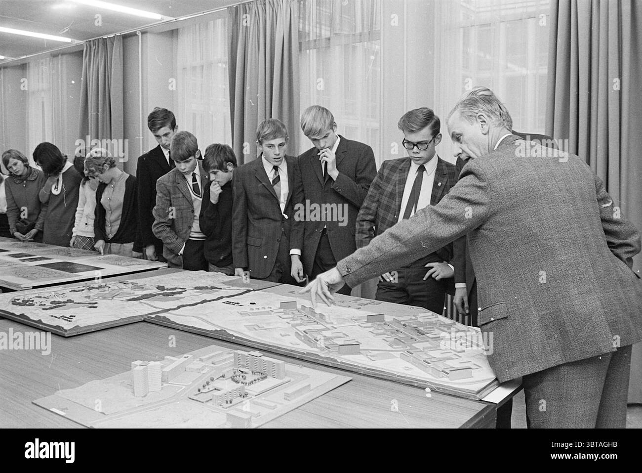 Construction Day IJmuiden Construction Site Meetings, Whizgle News, Dutch Desk, Niederlande, 1950 - 2000 am 08-11-1968. Diese Themen sind in der Abbildung dargestellt. Die Szene zeigt eine Zusammenkunft von Einzelpersonen um einen langen Tisch mit detaillierten architektonischen Modellen und Plänen, die wahrscheinlich ein geplantes Entwicklungs- oder Stadtprojekt darstellen. Der Tisch ist mit verschiedenen topografischen Darstellungen gefüllt, von denen einige komplexe Gebäudelayouts und Wege darstellen. Im Vordergrund steht ein älterer Mann, gekleidet in einem maßgeschneiderten grauen Anzug, auf eines der Models, vermutlich Ex Stockfoto