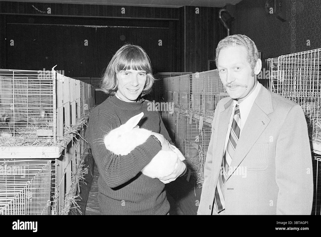 Kleine Tierausstellung S'poort Animals, Whizgle News, Dutch Desk, Niederlande, 1950 - 2000 am 05-11-1976. Die Abbildung zeigt diese Themen. Die Szene zeigt zwei Männer in einem Innenbereich, wahrscheinlich eine Einrichtung, die mit Tierpflege oder Forschung zu tun hat. Ein Individuum mit langen, fließenden Haaren hält ein großes weißes Kaninchen nahe an der Brust und zeigt eine sanfte Interaktion zwischen Mensch und Tier. Das Kaninchen ist flauschig und wirkt ruhig, was dem Moment einen warmen, liebevollen Ton verleiht. Der zweite Mann, der neben dem ersten steht, trägt einen formellen Anzug, der im Kontrast zur Freizeitkleidung steht Stockfoto