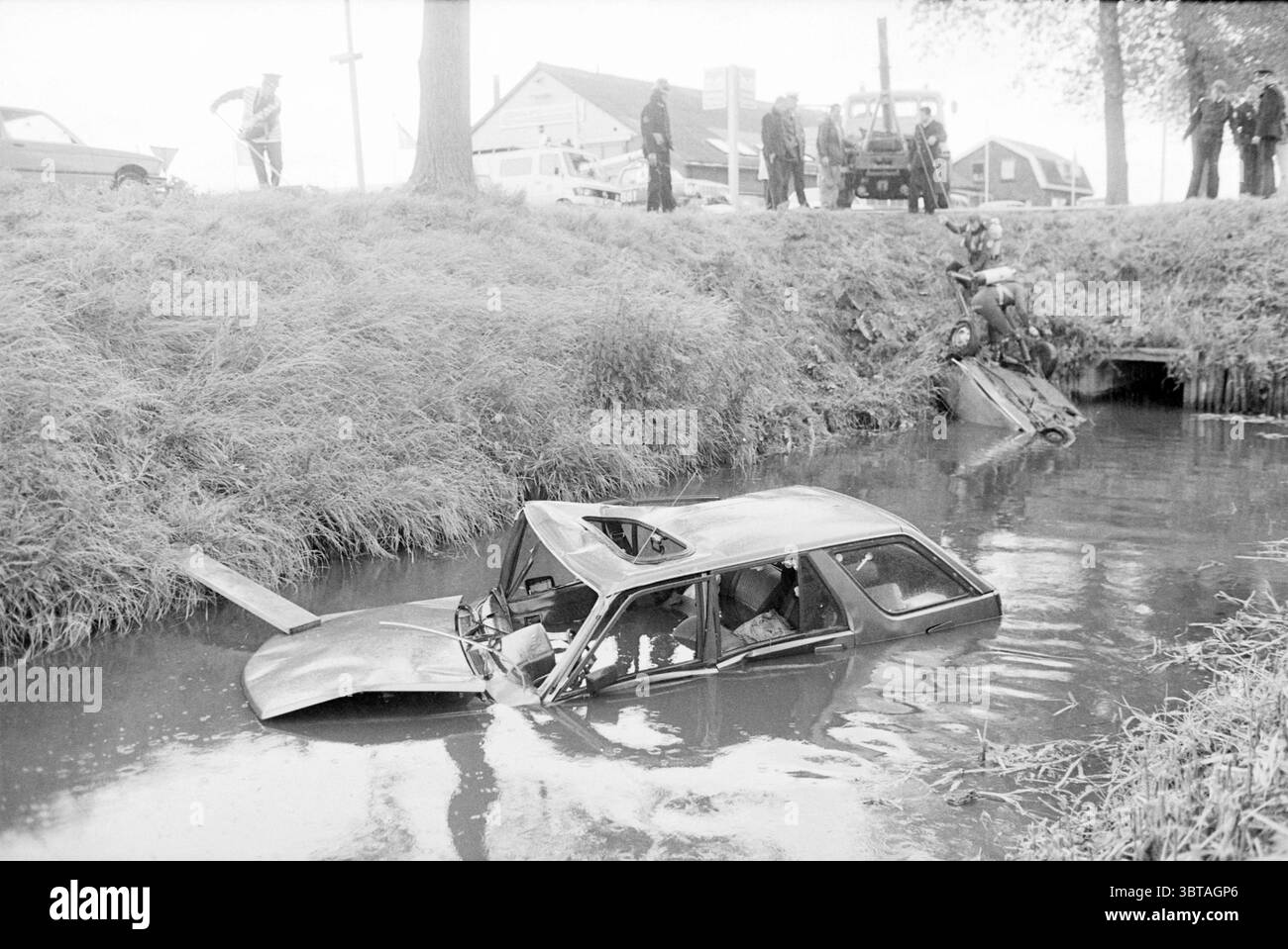 Autos im Wasser nach einer Kollision., Whizgle News, Dutch Desk, Niederlande, 1950 - 2000 am 07-1984. Diese Themen werden in der Abbildung angezeigt. Die Szene zeigt einen düsteren und dramatischen Moment nach einem Fahrzeugunfall. Im Vordergrund ist ein Auto teilweise in einen trüben Wasserkörper getaucht, dessen Dach leicht über der Oberfläche liegt. Das Fahrzeug scheint beschädigt zu sein, mit zerknittertem Metall und zersplitterten Fenstern, was auf einen heftigen Zusammenstoß hindeutet. Das Wasser um das Auto herum reflektiert einen grauen Ton und verschmilzt mit den dunklen Farben des bewölkten Himmels, was die ernste Stimmung der Szene noch verstärkt. Hohes Gras und Unkraut Stockfoto