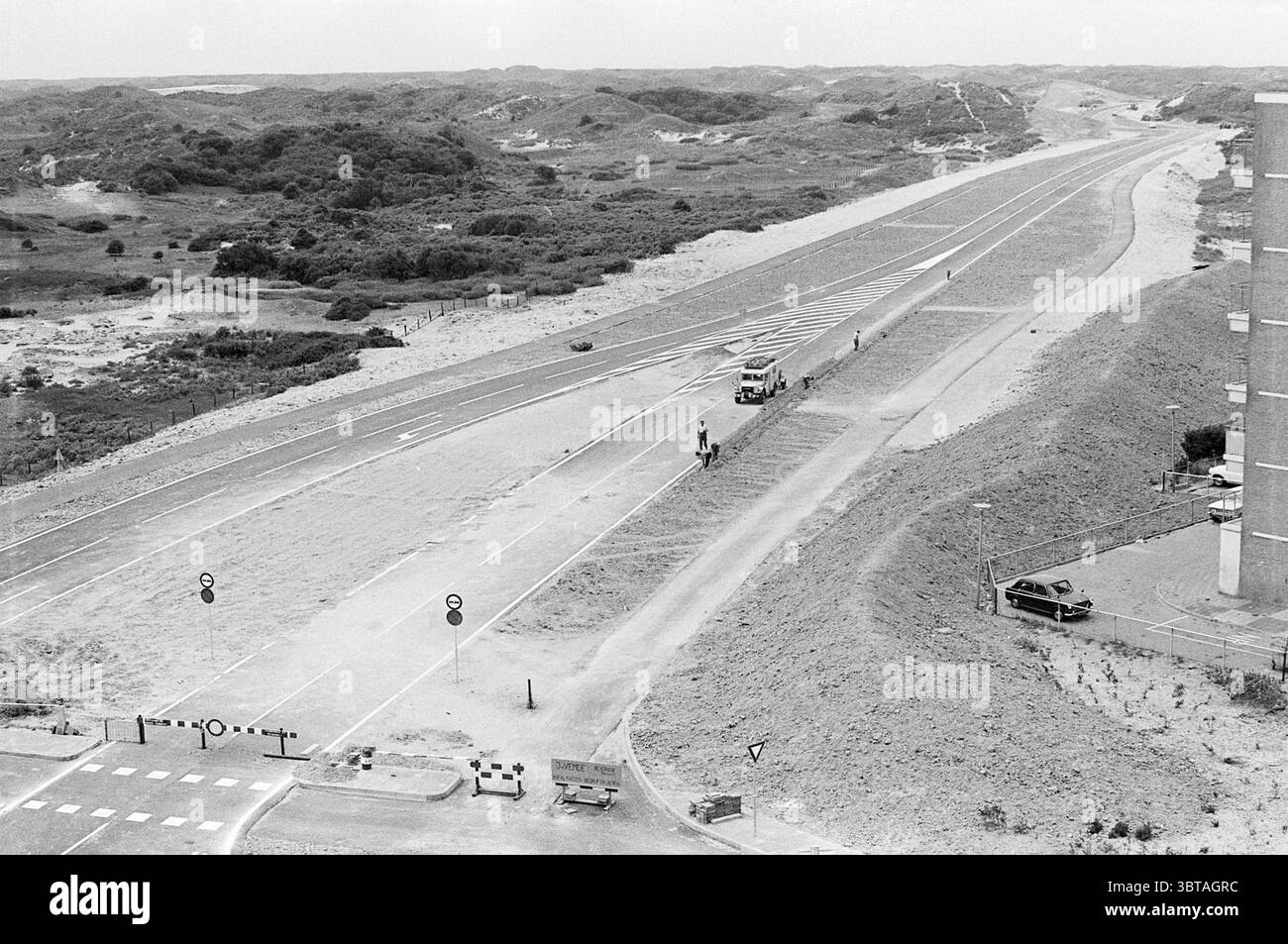 Arbeiten Sie an der Verlängerung des Herenduinweges. Work, Whizgle News, Dutch Desk, Niederlande, 1950 - 2000 am 25-06-1973. Diese Themen sind in der Abbildung dargestellt. Die Szene zeigt eine weite, offene Landschaft, die von einer weiten, leeren Straße dominiert wird, die sich bis in die Ferne erstreckt. Die Straße wird von Sandflächen auf beiden Seiten flankiert und ist durchsetzt von dünnen Vegetationsflächen, einschließlich Grasklumpen und niedrigen Sträuchern. Die Umgebung zeichnet sich durch sanfte, hügelige Hügel aus, die sich in den Hintergrund zurückziehen und ein Gefühl von Tiefe erzeugen. Die Atmosphäre hat einen gedämpften, monochromatischen Ton, wahrscheinlich aufgrund der Wahl Stockfoto