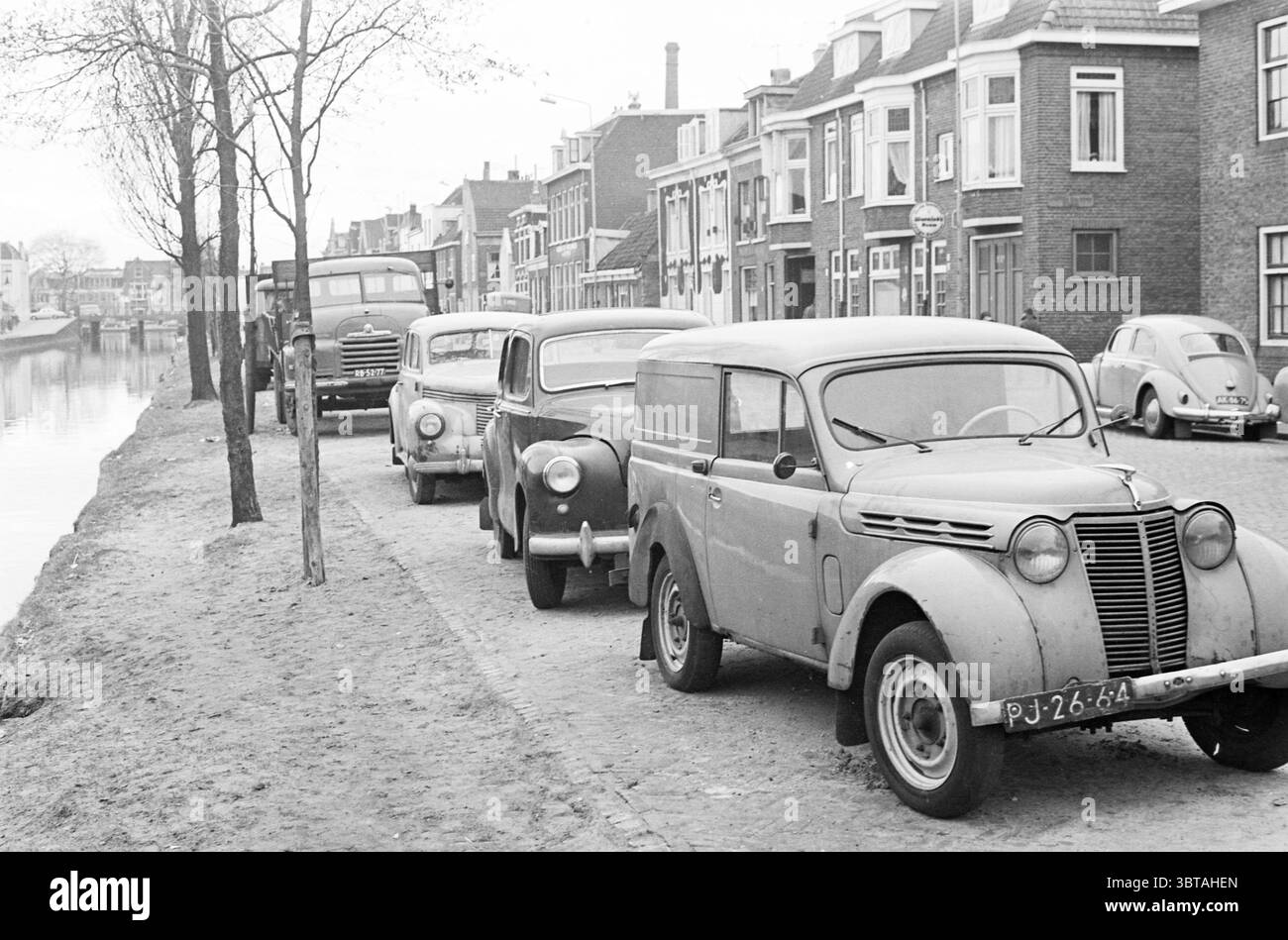 Parkchaos entlang lange Herenvest und Herensingel Haarlem lange Herenvest Nederland., Whizgle News, Dutch Desk, Niederlande, 1950 - 2000. Das Bild enthält diese Themen. Die Szene zeigt eine malerische urbane Umgebung entlang eines engen Kanals, der von alten Häusern gesäumt ist. Auf der einen Seite parken mehrere Oldtimer dicht nebeneinander auf einer kopfsteingepflasterten Straße, deren verwitterte Oberflächen darauf hindeuten, dass sie bessere Tage gesehen haben. Die Farben sind überwiegend gedämpft, wobei Grau- und Blautöne die Fahrzeuge und Gebäude dominieren. Die Fahrzeuge unterscheiden sich im Stil und zeigen Designs aus vergangenen Zeiten mit abgerundeter Kante Stockfoto