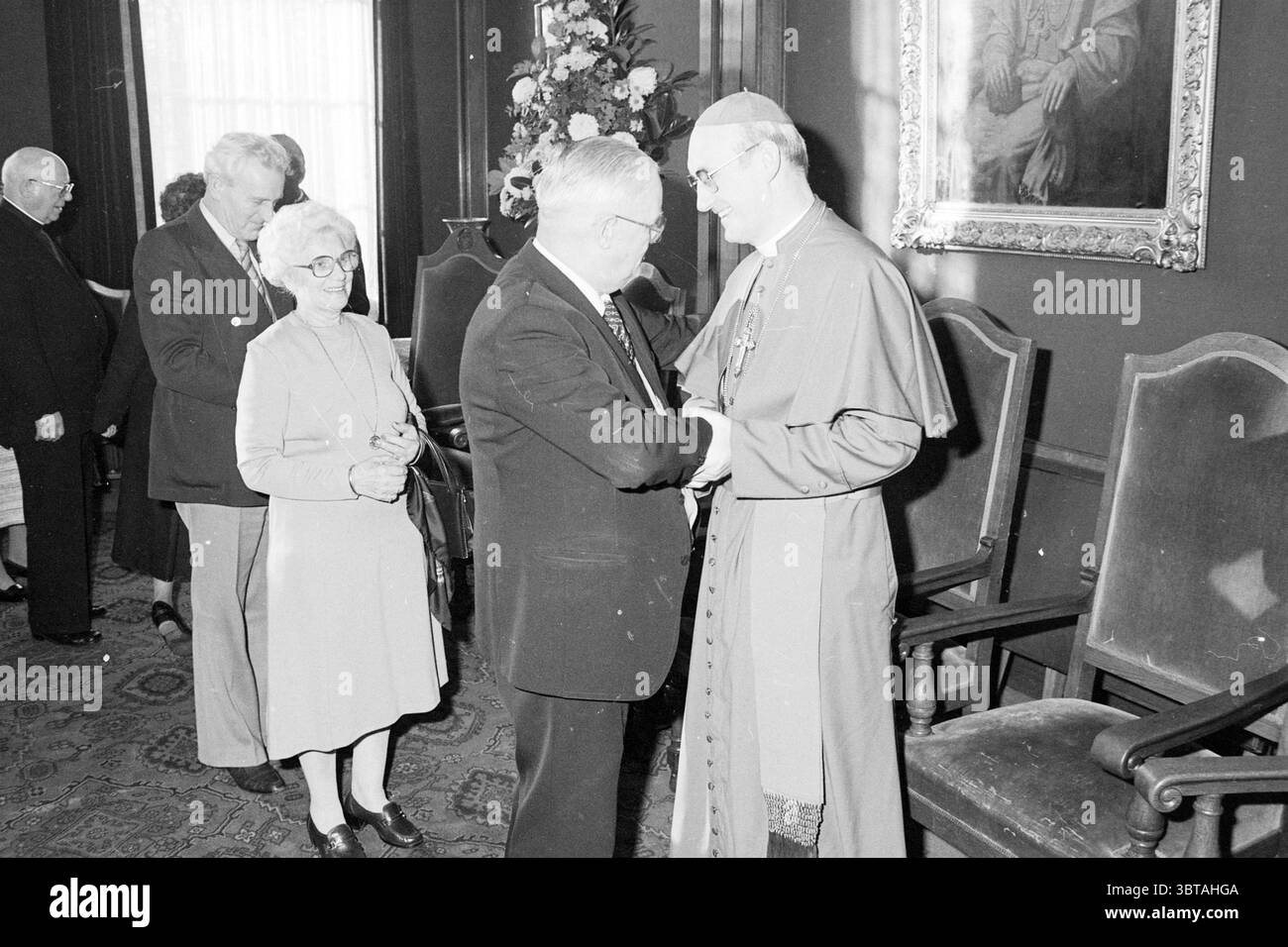 Ordination von Bischof Bomers und Weihbischof Lescrauwaert RK Bavo Haarlem Weihe römisch-katholische Kirche Haarlem Niederlande, Whizgle News, Dutch Desk, Niederlande, 1950 - 2000 am 14-01-1984. Dies sind die Themen im Bild. In einem warm beleuchteten Raum findet eine Versammlung von mehreren Personen statt, wahrscheinlich in einer formellen Umgebung. Die Wände sind in satten Farbtönen gehalten, vielleicht in einem tiefen Burgunderrot oder Waldgrün, und große Fenster lassen das Tageslicht durchleuchten, das ein sanftes Leuchten über die Szene wirft. Im Vordergrund greift ein Mann in formeller Kleidung, einschließlich eines dunklen Anzugs und einer Krawatte, mich an Stockfoto