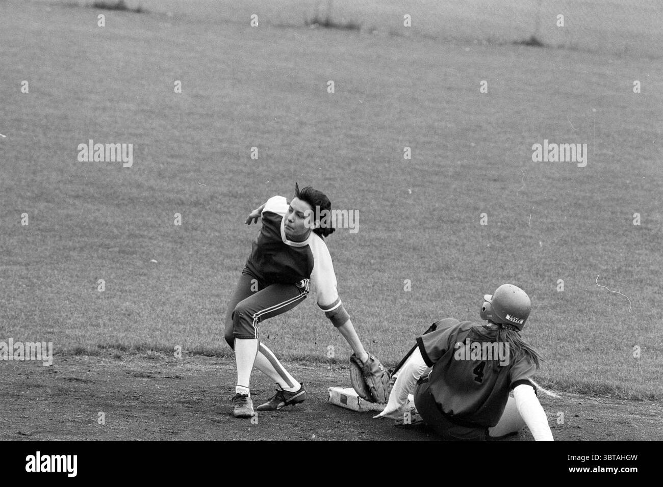 Softball Pioneers - HVV, Whizgle News, Dutch Desk, Niederlande, 1950 - 2000 am 08-06-1991. Diese Themen sind in der Abbildung dargestellt. In einer dynamischen Szene auf einem Softballfeld spielen zwei Spieler einen entscheidenden Moment. Ein Athlet in einem weiß-tiefblauen Trikot mit passender Hose scheint in Bewegung zu sein, nachdem er gerade die Basis markiert hat. Sie haben dunkles, gewelltes Haar, das hinter ihnen fließt und ein Gefühl von Dringlichkeit und Handlung vermittelt. Der andere Spieler in einem dunkelfarbenen Trikot mit der Nummer 4 liegt auf dem Boden und scheint gerade die Basis erreicht zu haben. Sie trägt einen Helm, der rot erscheint, im Kontrast zu wi Stockfoto