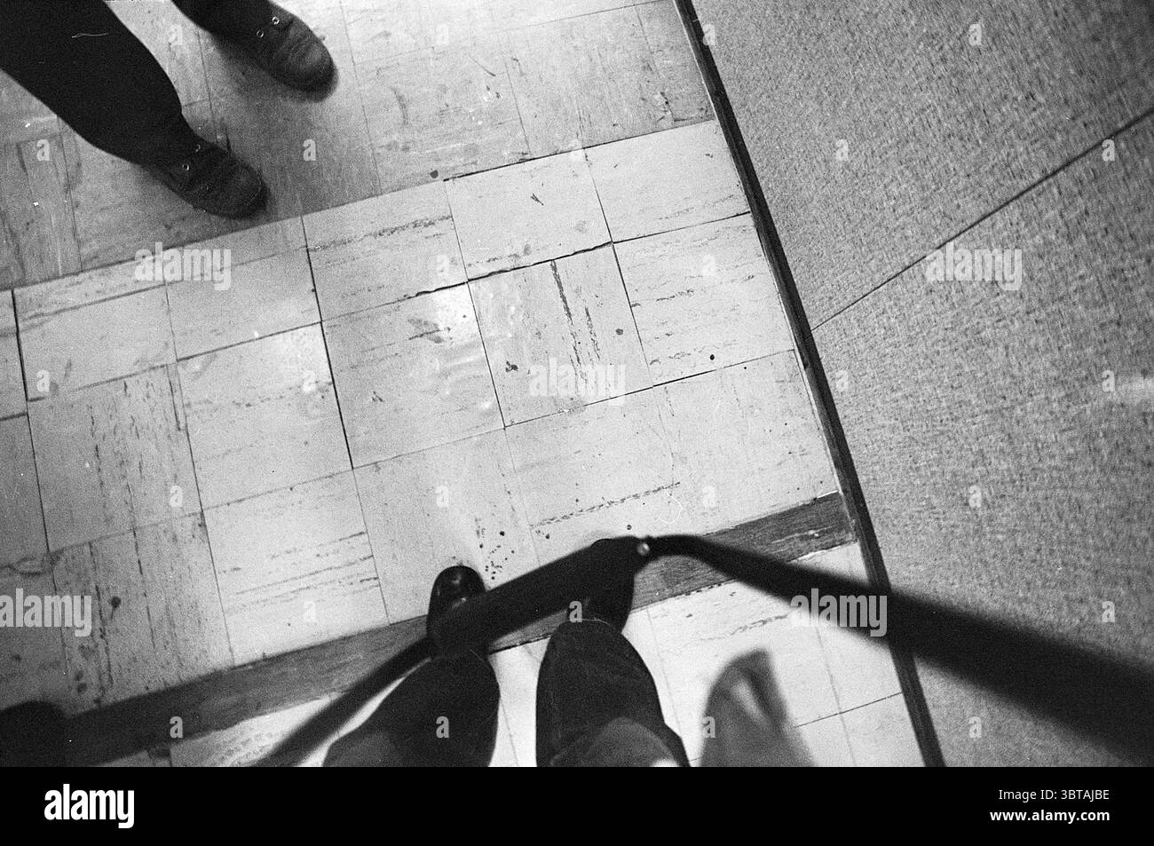 Unbreakable., Whizgle News, Dutch Desk, Niederlande, 1950 - 2000. Die Abbildung zeigt diese Themen. Die Szene zeigt eine Bodenoberfläche aus nächster Nähe und fängt eine Atmosphäre der Intimität und vielleicht ein Gefühl der Einsamkeit ein. Der Bodenbelag zeigt ein Muster aus hellen und dunklen Fliesen, wobei die hellen Töne einen leicht gedämpften, schwachen Glanz aufweisen, der sich von den dunkleren Bereichen abhebt. Diese Fliesen deuten auf ein Gefühl von Alter oder Verschleiß hin, was auf häufigen Fußverkehr hindeutet. Am unteren Rand des Rahmens befinden sich zwei Paar Füße, von denen einer in einem dunklen, strukturlosen Schuh, möglicherweise einem Schuh, gekleidet ist, während der andere Fuß getragen wird Stockfoto