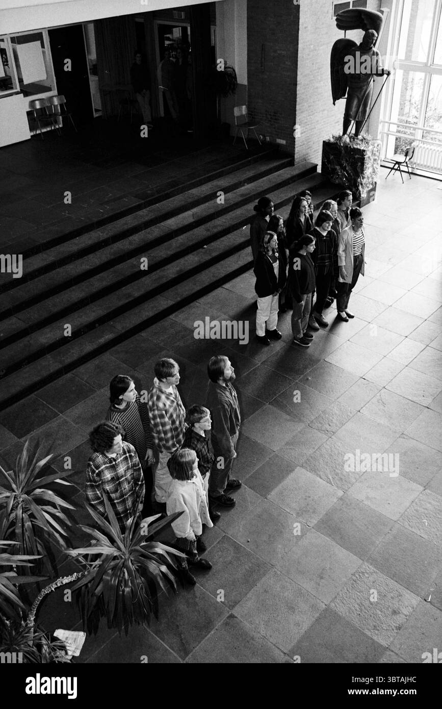 Theaterprobe Santa Maria, Whizgle News, Dutch Desk, Niederlande, 1950 - 2000 am 11-03-1997. Das Bild enthält diese Themen. Die Szene spielt in einer geräumigen Innenumgebung, die durch große Steinplatten gekennzeichnet ist, die ein Gefühl von Beständigkeit und Struktur schaffen. Die allgemeine Atmosphäre fühlt sich besinnlich an, vielleicht sogar etwas feierlich, verstärkt durch die monochromatische Farbpalette von Schwarz und weiß. Im Vordergrund steht eine Gruppe von Individuen in einer einzigen, geordneten Linie, mit Blick auf eine überlebensgroße, plastische Figur, die Stärke oder Wachsamkeit zu symbolisieren scheint. Die Gruppe besteht aus o Stockfoto