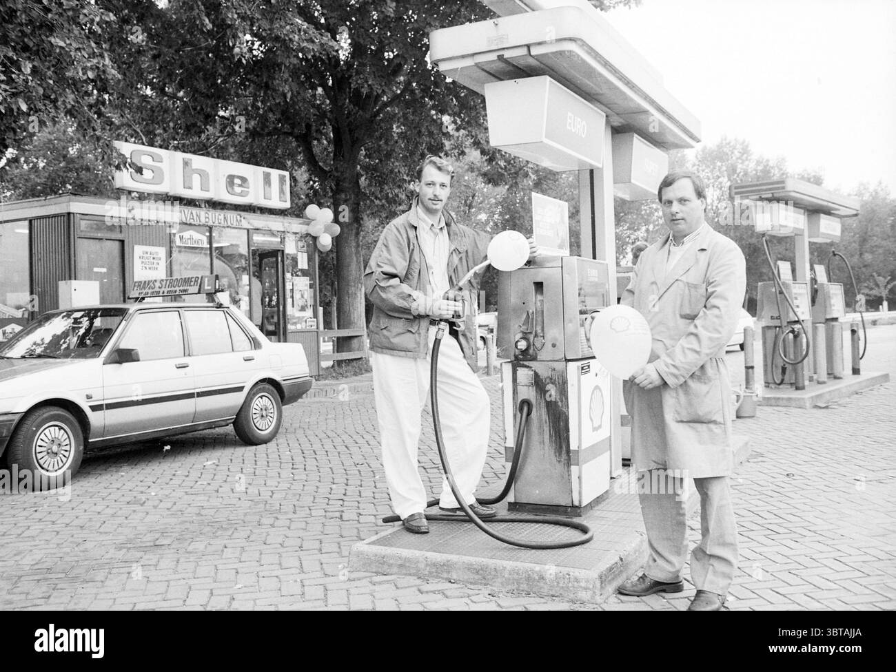 Zwei Männer in der Shell Van Bugnum Pumpstation in Santpoort Santpoort, Whizgle News, Dutch Desk, Niederlande, 1950 - 2000 am 02-10-1989. Dies sind die Themen im Bild. In einer belebten Tankstelle stehen zwei Männer neben einer altmodischen Kraftstoffpumpe. Beide tragen helle Outfits, von denen eines in einer lockeren Jacke und das andere in einem langen Mantel trägt. Ihre Ausdrücke scheinen entspannt, aber engagiert zu sein, was ein Gefühl von Kameradschaft erzeugt, während sie weiße Gaspumpendüsen halten und sich wahrscheinlich darauf vorbereiten, ein Fahrzeug zu betanken. Der Hintergrund zeigt eine Tankstellenstruktur mit einem ausgeprägten Stil, teilweise undurchsichtig Stockfoto