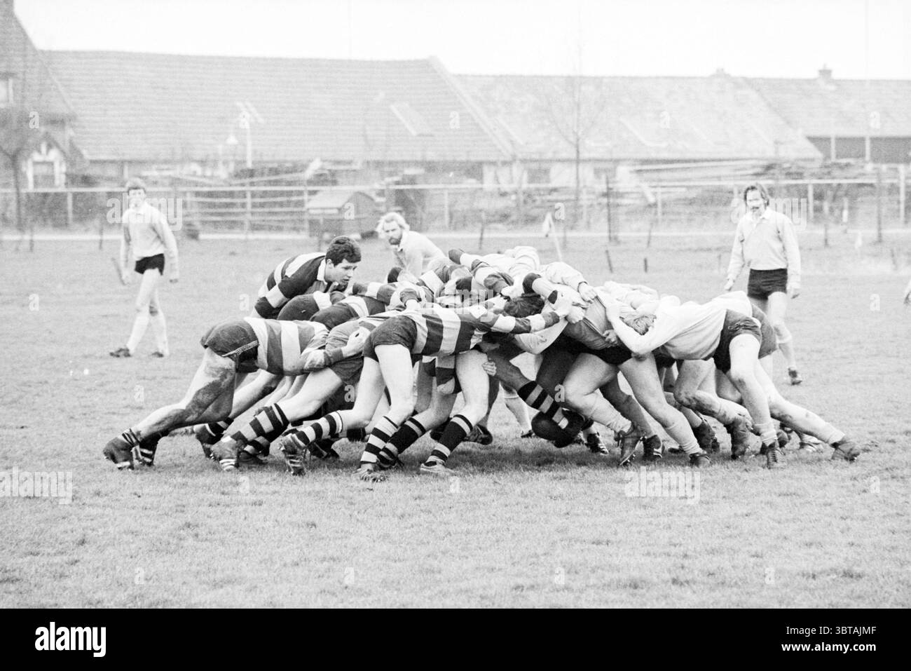 Smugglers - Castricum Rugby, Whizgle News, Dutch Desk, Niederlande, 1950 - 2000 am 09-03-1980. Dies sind die Elemente im Bild. Auf einem von Feuchtigkeit gekennzeichneten grasbewachsenen Feld spielen zwei Teams einen dynamischen Rugby-Scrum. Die Spieler, die in gestreiften Trikots – einige in dunklen Farben, andere in helleren Tönen – gekleidet sind, bilden eine eng gepackte Gruppe, deren Körper sich mit intensiver Anstrengung ineinander lehnen. Der Kontrast der Uniformen erzeugt einen Rhythmus aus schwarzen und weißen Streifen, der ihre kollektive Stärke in diesem entscheidenden Moment des Spiels unterstreicht. Die Umgebung bietet eine Kulisse aus bescheidenen, niedrigen Gebäuden mit Stockfoto