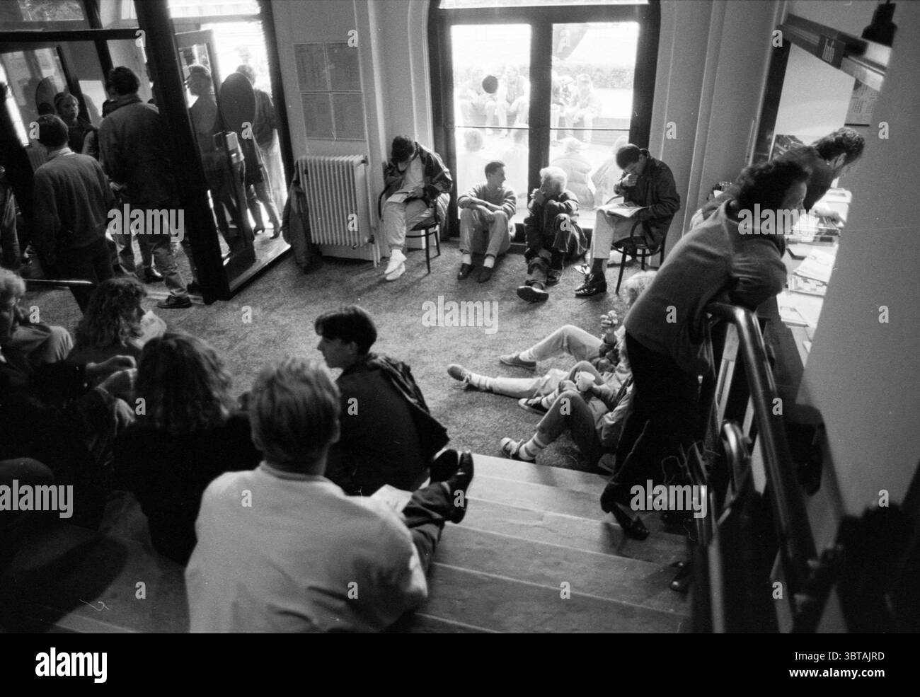 Geschäftige Einzelverkäufe Stadsschouwburg Haarlem Haarlem Wilsonsplein 23 The Netherlands, Whizgle News, Dutch Desk, Niederlande, 1950 - 2000 am 06-09-1991. Dies sind die Elemente im Bild. Die Szene fängt einen belebten, intimen Versammlungsraum ein, der auf einen Moment des Übergangs oder einer Pause hinweist. Eine vielfältige Gruppe von Menschen bevölkert den Raum, einige sitzen auf Holzstühlen, während andere auf dem Boden verstreut sind, in Gedanken oder Gesprächen verloren. Die gesamte Farbpalette ist gedämpft, dominiert von Grau- und Schwarztönen, was eine zeitlose, nostalgische Qualität verleiht. Der Boden ist mit Teppichboden ausgelegt, was die Atmosphäre weicher macht Stockfoto