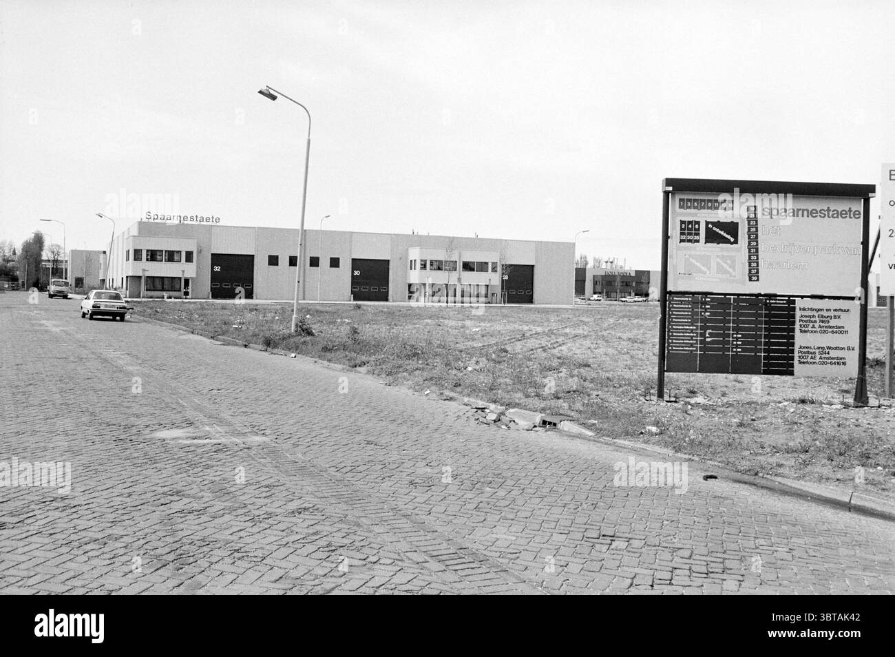 Waarderpolder Piloten Haarlem the Netherlands, Whizgle News, Dutch Desk, Niederlande, 1950 - 2000 am 02-05-1983. Das Bild enthält diese Themen. In der Szene erstreckt sich eine breite, offene Straße zum Horizont hin, flankiert von einem Grasfleck auf der einen Seite und einem Industriebau auf der anderen Seite. Die Struktur, die mit einer modernen Ästhetik gestaltet wurde, verfügt über eine Kombination aus großen Fenstern und flachen Oberflächen, die in neutralen Grau- und Weißtönen gestrichen sind. Die Architektur des Gebäudes verleiht ihm ein klares, minimalistisches Aussehen, mit mehreren Abschnitten, die verschiedene Funktionen oder o anzeigen Stockfoto