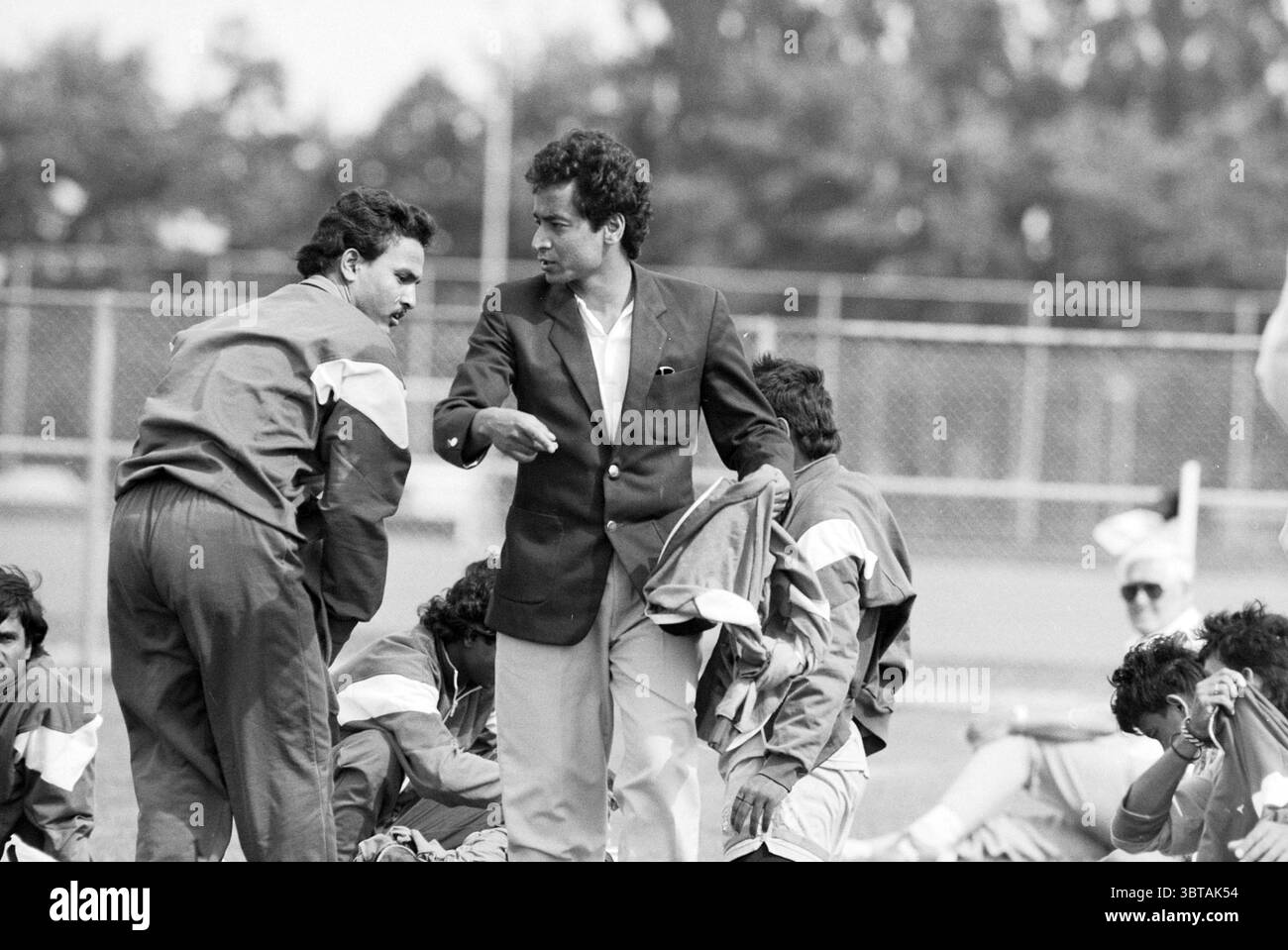 Hollandia - Indien + Regisseur + Coach India, Whizgle News, Dutch Desk, Niederlande, 1950 - 2000 am 08-08-1991. Das Bild enthält diese Themen. In einer dynamischen Umgebung im Freien versammelt sich eine Gruppe von Individuen auf einem grasbewachsenen Feld, die wahrscheinlich eine sportliche Aktivität ausüben. Die Szene ist durch eine monochromatische Palette gekennzeichnet, die ihr ein klassisches Gefühl verleiht, das an frühere Epochen erinnert. Im Mittelpunkt der Komposition stehen zwei Männer, die sich unterhalten. Einer trägt einen formellen Anzug mit Blazer, ein auffälliger Kontrast zu den anderen sportlichen Freizeitbekleidung, die aus aufeinander abgestimmten Trainingsanzügen besteht. Diese Atti Stockfoto
