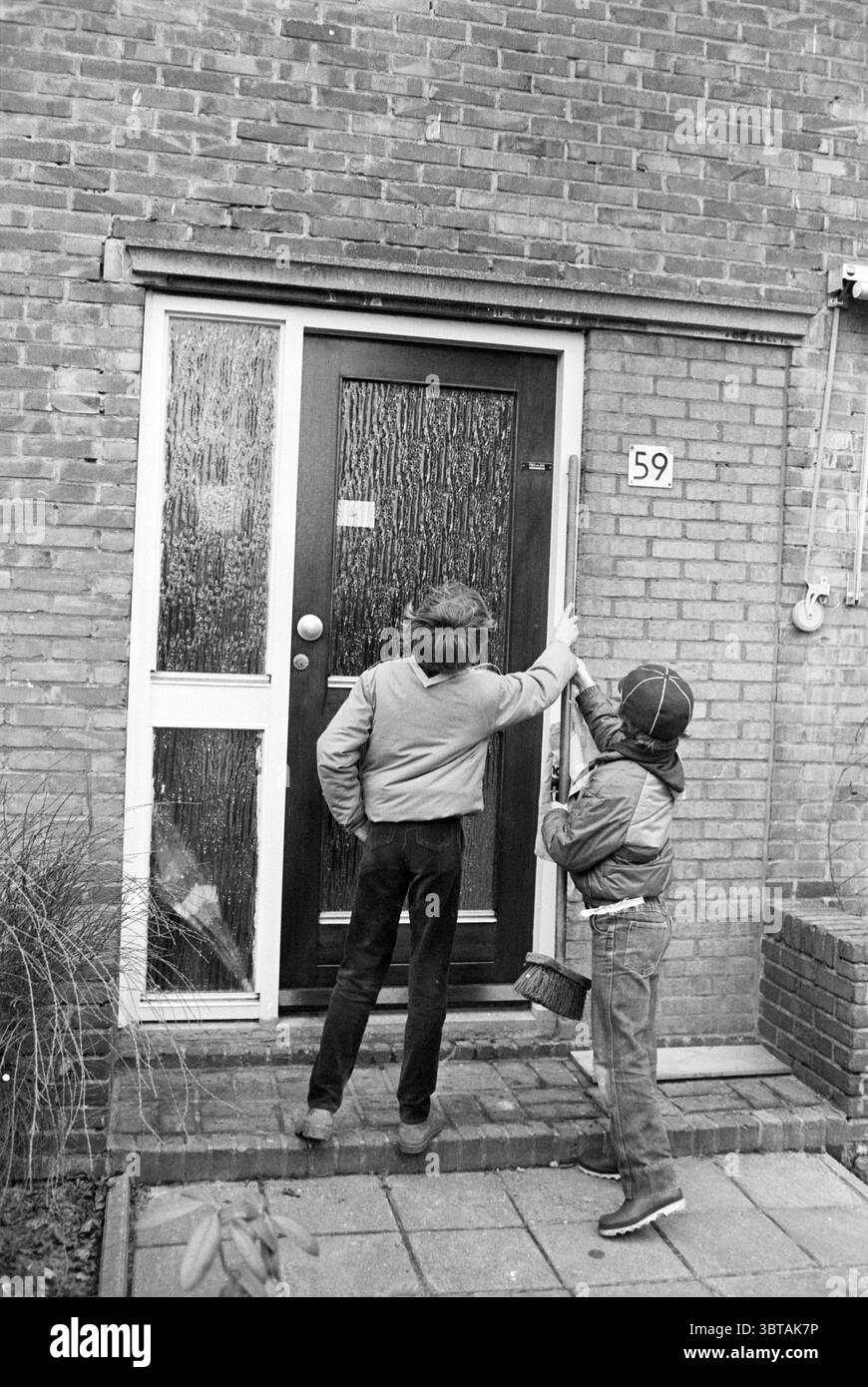 Junge Pfadfinder reinigen einen Park und Gehweg, Whizgle News, Dutch Desk, Niederlande, 1950 - 2000 am 11-04-1985. Dies sind die Elemente im Bild. In einer gedeckten, monochromen Umgebung stehen zwei Kinder vor einer Ziegelwand und sehen eine Tür. Die Ziegel haben einen tiefen Schatten, der der Szene Textur und Tiefe verleiht. Die Kinder, beide in für diese Zeit typischen Freizeitkleidung gekleidet, scheinen konzentriert zu sein. Ein Kind mit zerzausten Haaren und einer Jacke reicht zur Tür, während das andere, etwas kleiner und mit einer Mütze bekleidet, aufmerksam beobachtet. Die Tür selbst ist massiv und hölzerne, wie angestrichen Stockfoto