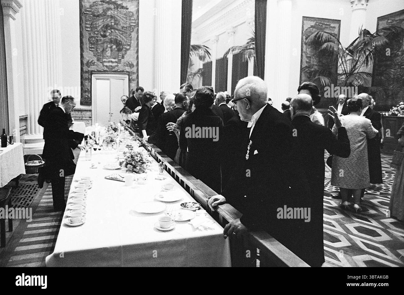 Prinzessin Margriet beim Galakonzert 150 Jahre N.P.O. königliche Empfänge und königliche Besuche des North Holland Philharmonic Orchestra, Whizgle News, Dutch Desk, Niederlande, 1950 - 2000 am 29.11.1963. Dies sind die Themen im Bild. Die Szene fängt ein formelles Treffen in einem eleganten Saal ein, gekennzeichnet durch hohe Decken mit komplizierten Verzierungen. Die Wände sind mit großen Wandgemälden dekoriert, die Szenen aus Natur und Kultur darstellen und der ansonsten gedämpften Atmosphäre einen Hauch von Lebendigkeit verleihen. An der Vorderseite erstreckt sich ein langer Banketttisch über den Raum, der mit einem weißen Tisch bedeckt ist Stockfoto