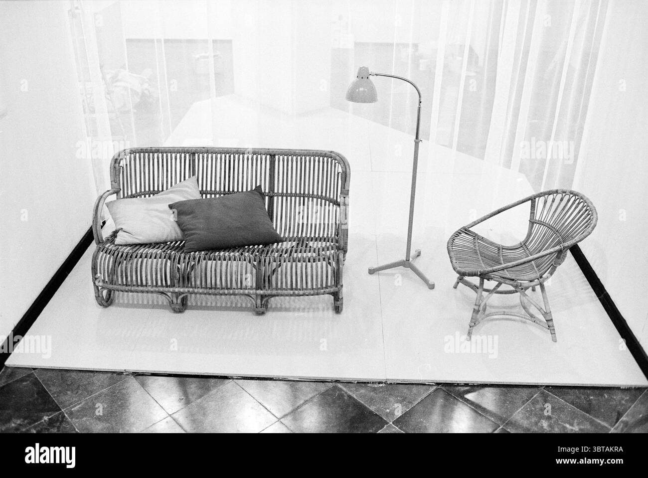 Möbelanordnung in der Ausstellung „Good Living“ in Vleeshal Haarlem Niederlande, Whizgle News, Dutch Desk, Niederlande, 1950 - 2000 am 01-03-1979. Das Bild enthält diese Themen. In einer minimalistischen Inneneinrichtung steht ein Rattan-Sofa im Mittelpunkt, dessen geschwungene Linien und gewebte Textur ein Gefühl natürlicher Eleganz ausstrahlen. Das Sofa ist mit einigen dekorativen Kissen in weichen, gedämpften Farben verziert, die einen Hauch von Komfort und Wärme verleihen. Neben dem Sofa befindet sich eine schlanke Stehleuchte, deren klare Linien und die Metallic-Oberfläche einen starken Kontrast zum organischen Gefühl des Rattans bilden Stockfoto