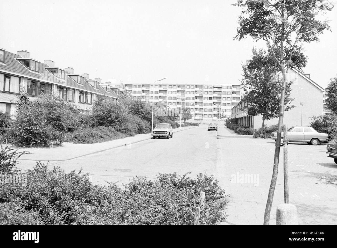 Haarlemmermeer 1980 Haarlemmermeer Haarlemmermeer, Whizgle News, Dutch Desk, Niederlande, 1950 - 2000 am 01-08-1980. Dies sind die Elemente im Bild. Die Szene präsentiert ein ruhiges Wohnviertel, das durch eine Mischung aus zweistöckigen Häusern und mehrstöckigen Wohngebäuden gekennzeichnet ist. Auf der linken Seite säumen mehrere Häuser die Straße, mit schrägen Dächern und sanften Tönen, die ein Gefühl von Wärme hervorrufen. Sie sind teilweise von üppigen grünen Büschen verdeckt, die der städtischen Umgebung ein natürliches Element verleihen. Auf der anderen Straßenseite erstreckt sich ein moderner Block von Appartements in die Ferne, Showcas Stockfoto