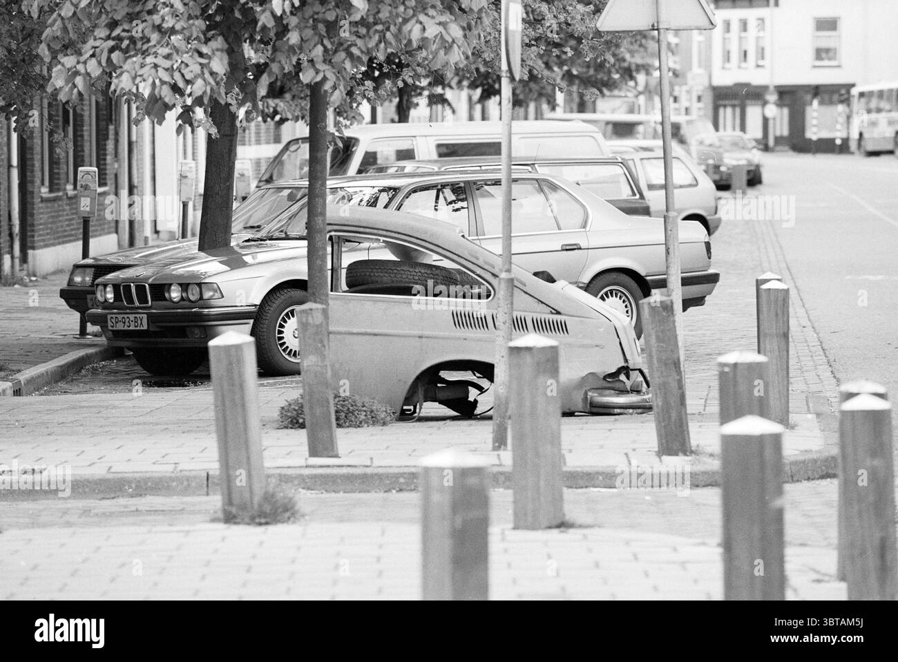 Ein halbes Auto auf dem Bürgersteig in Nassaulaan Haarlem Nassaulaan Niederlande, Whizgle News, Dutch Desk, Niederlande, 1950 - 2000 am 15.05.1991. Das Bild enthält diese Themen. Die Szene zeigt eine ruhige Stadtstraße, die hauptsächlich durch eine Mischung aus geparkten Autos und einem Gefühl der Verlassenheit gekennzeichnet ist. Im Vordergrund befindet sich ein teilweise demontiertes Fahrzeug, das wie ein älteres Modell aussieht und auf einer Kopfsteinoberfläche aufliegt. Der Rahmen ist abgestutzt und zeigt Mechanismen und Teile, die sich in starkem Kontrast zur Umgebung befinden. Die Struktur des Fahrzeugs ist metallisch und langweilig, was darauf hindeutet Stockfoto