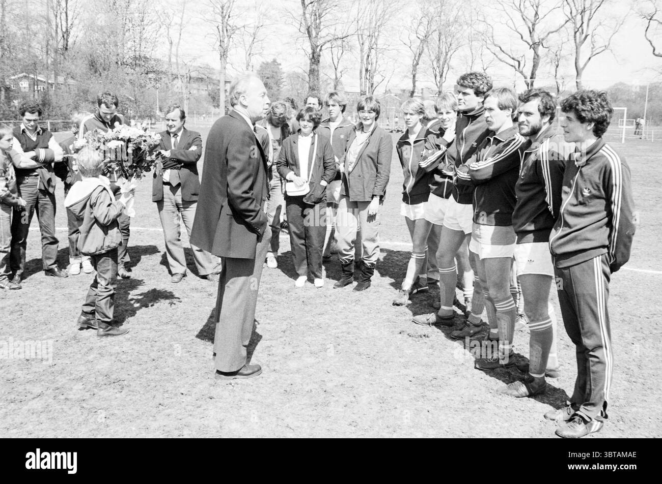 Zu Ehren der Bloemendaal-Spieler (Fußball) Willemsen und Van der Putten Football People and Trainer, Whizgle News, Dutch Desk, Niederlande, 1950 - 2000 am 25-04-1982. Dies sind die Elemente im Bild. Die Szene spielt in einem grasbewachsenen Gebiet, wahrscheinlich einem Sportplatz, unter hellem Tageslicht mit klarem Himmel. Das Sonnenlicht schafft eine warme, einladende Atmosphäre. Eine Gruppe von Menschen, die sowohl Männer als auch Frauen umfasst, bildet einen Halbkreis, der sich um einen älteren Mann versammelt, der vor ihnen steht. Der ältere Mann, in einem dunklen Anzug gekleidet, führt Gespräche, seine Gesten zeigen ein Gefühl von Autorität oder Stockfoto