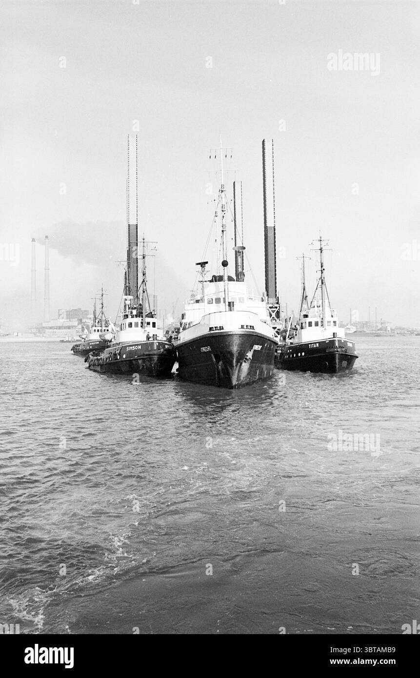 Abschleppen von Bohrplattformteilen, Whizgle News, Dutch Desk, Niederlande, 1950 - 2000 im Jahr 1967. Dies sind die Themen im Bild. Die Szene zeigt eine Gruppe von Booten, die eng aneinander auf dem Wasser ankern, deren Rümpfe in verschiedenen Schwarz-weiß-Tönen gemalt sind und eine klassische maritime Ästhetik widerspiegeln. Das mittlere Schiff ist größer, mit einer stabilen Struktur und einer markanten Takelage, während zwei kleinere Boote, die es flankieren, stromlinienförmiger erscheinen, was darauf hindeutet, dass sie für Manövrierfähigkeit ausgelegt sind. Der Himmel darüber ist ein gedämpftes Grau, was eine düstere Stimmung schafft, die im Kontrast zur dunkleren Präsenz von steht Stockfoto
