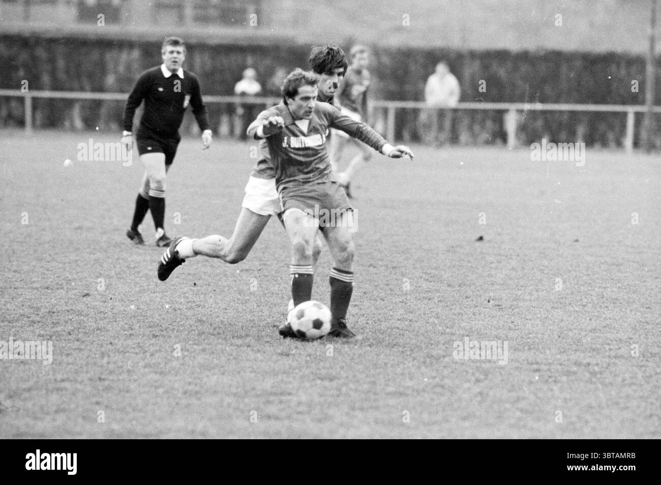 Fulmars - DRC Football, Whizgle News, Dutch Desk, Niederlande, 1950 - 2000 am 09-01-1983. Diese Themen werden in der Abbildung angezeigt. Die Szene fängt einen intensiven Moment auf einem grasbewachsenen Fußballfeld ein. Zwei Spieler kämpfen um den Besitz eines auf dem Boden liegenden Fußballs. Ein Spieler, der in ein tiefblaues Set gekleidet ist, scheint etwas voraus zu sein und sich entschlossen nach vorne zu lehnen. Sein Gesicht drückt Fokus und Körnung aus, während seine Beine dynamisch positioniert sind, als wäre er bereit zum Sprinten oder Drehen. Der gegnerische Spieler, in weiß gekleidet, ist dicht hinter ihm und bemüht sich, ihn abzufangen. Er ist sli Stockfoto