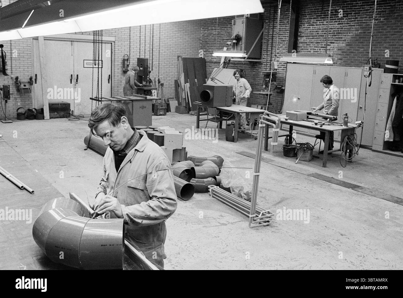 Werkstatt des PVC-Verarbeitungsunternehmens in IJmuiden IJmuiden Niederlande, Whizgle News, Dutch Desk, Niederlande, 1950 - 2000 am 28-11-1979. Dies sind die Themen im Bild. Die Szene entfaltet sich in einer geräumigen industriellen Werkstatt, die sich durch ihre hohe Decke und den weitläufigen Betonboden auszeichnet. Die Wände haben ein strukturiertes Aussehen, wahrscheinlich aus Ziegeln, was zu dem rauen, zweckmäßigen Ambiente beiträgt. Auf der linken Seite konzentriert sich ein Mann sehr darauf, ein großes, geschwungenes Metallstück zu Formen. Er trägt eine helle Arbeitsjacke, und die Konzentration auf seinem Gesicht deutet auf eine Aufgabe hin, die Präzision erfordert Stockfoto