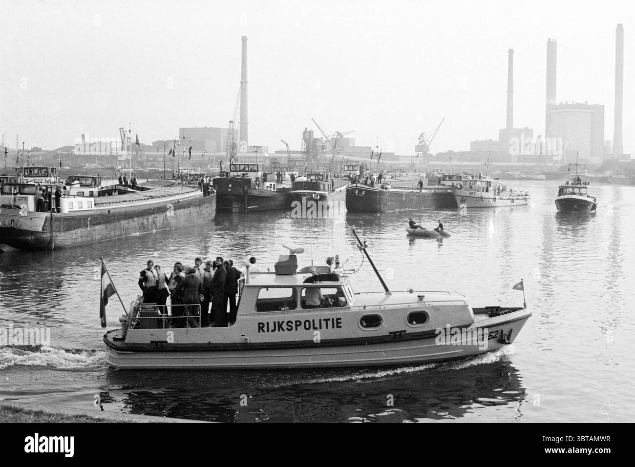 Beseitigung der Blockade des Nordseekanals IJm. North Holland Dörfer usw. North Sea Canal IJmuiden the Netherlands, Whizgle News, Dutch Desk, Niederlande, 1950 - 2000 am 11-08-1981. Das Bild enthält diese Themen. Die Szene zeigt einen belebten Hafen, an dem mehrere Boote angedockt sind, umgeben von einer Mischung aus Industriebauten und Maschinen. Im Vordergrund befindet sich ein Polizeiboot, das mit einer Flagge geschmückt ist, in Bewegung, mit einer Gruppe von Personen, die scheinbar an einer Diskussion oder Beobachtung beteiligt sind, was ein Gefühl von Autorität und Wachsamkeit suggeriert. Die Gruppe auf dem Polizeiboot ist in verschiedene o gekleidet Stockfoto