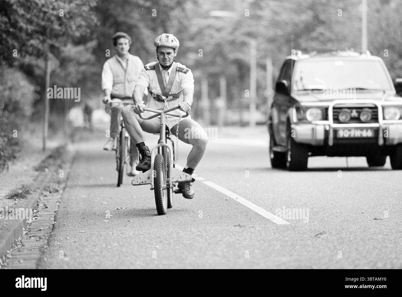 Witz mit Polizist Wagenweg Haarlem Wagenweg the Netherlands, Whizgle News, Dutch Desk, Niederlande, 1950 - 2000 am 17.09.1992. Die Abbildung zeigt diese Themen. In ruhiger urbaner Umgebung fahren zwei Radfahrer auf einer ruhigen Straße, die von Grün umgeben ist. Die Szene ist in Schwarz-weiß gehalten und verleiht der Atmosphäre eine nostalgische und zeitlose Qualität. Im Vordergrund steht ein Radfahrer in einem unkonventionellen Outfit, das ein lässiges Oberteil mit kurzen Shorts kombiniert. Er trägt einen Helm und zeigt einen verspielten Ausdruck, der auf eine sorglose Haltung hinweist. Sein Fahrrad, klein und unverwechselbar, Kontrast Stockfoto