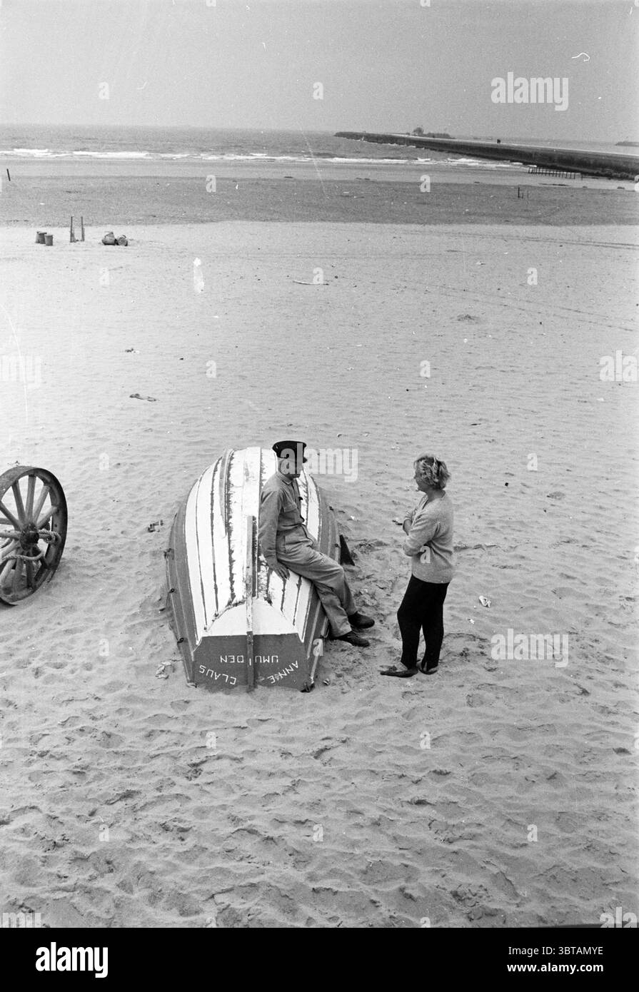 Beach Manager IJmuiden Persons Beach and Beaches, Whizgle News, Dutch Desk, Niederlande, 1950 - 2000 am 11-09-1961. Diese Themen sind in der Abbildung dargestellt. Die Szene entfaltet sich an einem Sandstrand, wo sich ein ruhiger Ozean im Hintergrund ausbreitet, dessen Wellen sanft am Ufer plätschern. Der Himmel darüber ist ein weiches, bedecktes Grau, das eine gedämpfte und ruhige Atmosphäre schafft. Im Vordergrund sitzt ein Mann auf einem verwitterten Holzboot, seine Haltung entspannt, während er sich zurücklehnt, scheinbar in Gedanken verloren. Er trägt legere Kleidung, die für den Strand geeignet ist, und einen Hut, der einen Hauch von Charakter verleiht. Sein Blick ist Stockfoto