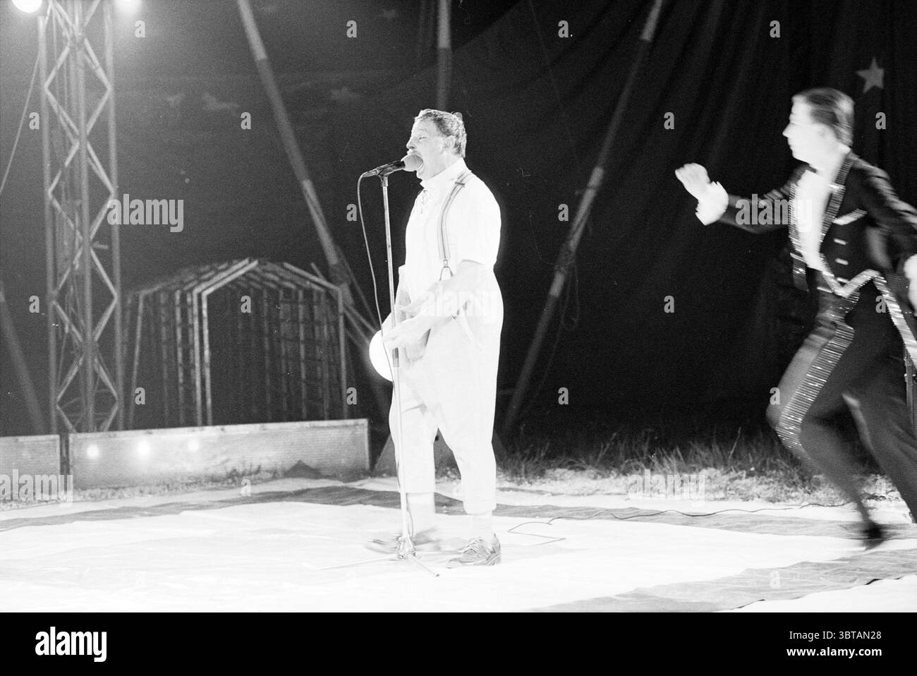 Circus Renz Schalkwijk Schalkwijk, Whizgle News, Dutch Desk, Niederlande, 1950 - 2000 am 08-05-1990. Das Bild enthält diese Themen. Die Szene zeigt eine fesselnde Zirkusvorstellung unter der schwachen Beleuchtung eines großen Zelt. In der Mitte steht ein Darsteller selbstbewusst an einem Mikrofon, das in einem weißen Outfit mit Hosenträgern gekleidet ist und ein Gefühl von Charisma ausstrahlt. Sein Ausdruck ist animiert, was darauf hindeutet, dass er das Publikum mit lebhaftem Geplänkel oder vielleicht einer komödiantischen Routine anspricht. Zu seiner Linken scheint ein anderer Künstler, der in einem eleganten schwarzen Anzug mit funkelnden Verzierungen gekleidet ist, bewegend zu sein Stockfoto